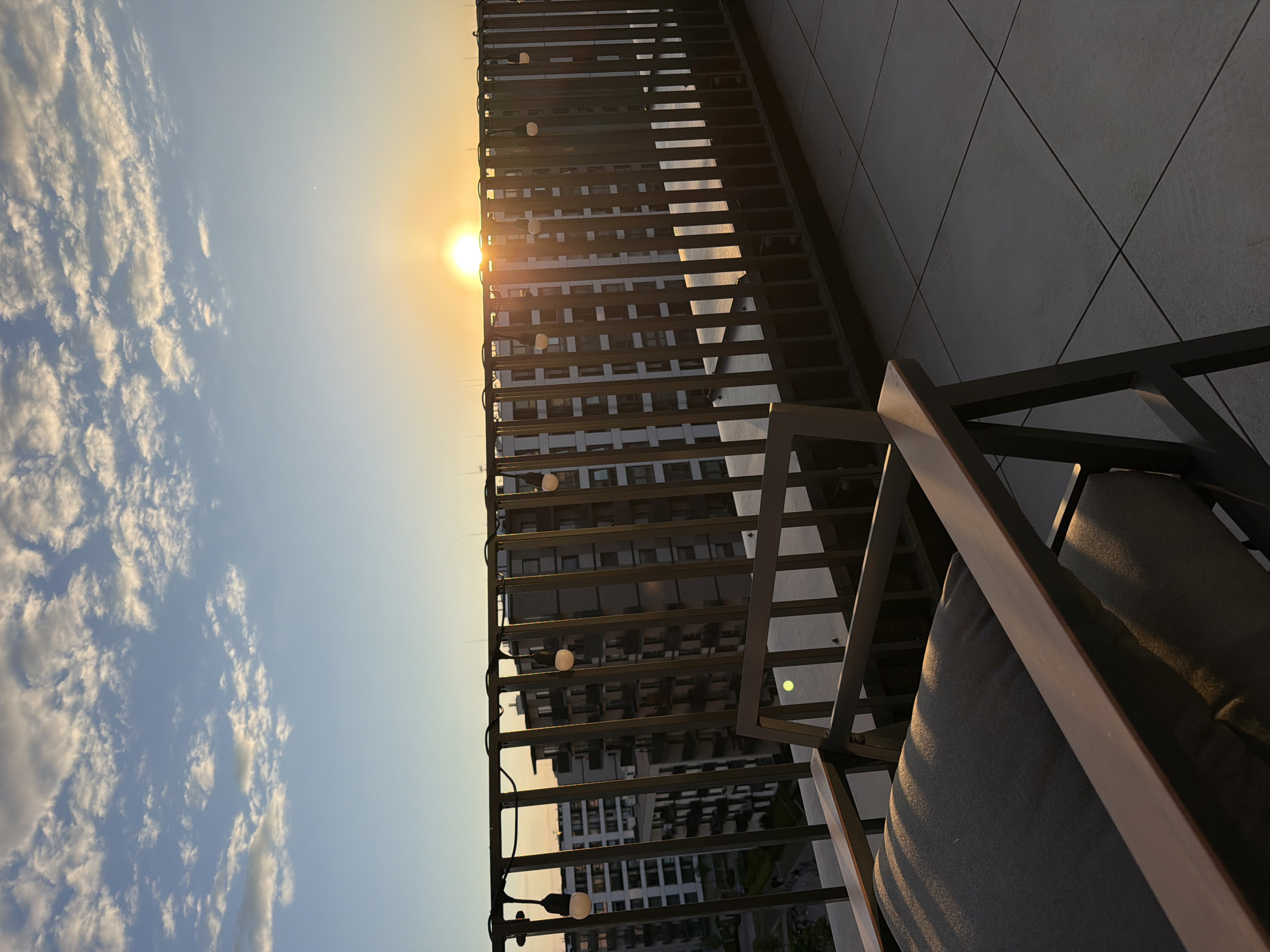 The image shows a view from a high-rise balcony at sunset. The balcony features a dark metal railing with string lights. A cushioned chair is partially visible in the lower right. The view beyond the railing includes a building with many windows, partially obscured by the railing and sun. The sky is blue with some clouds near the horizon. The location appears to be in an urban setting.