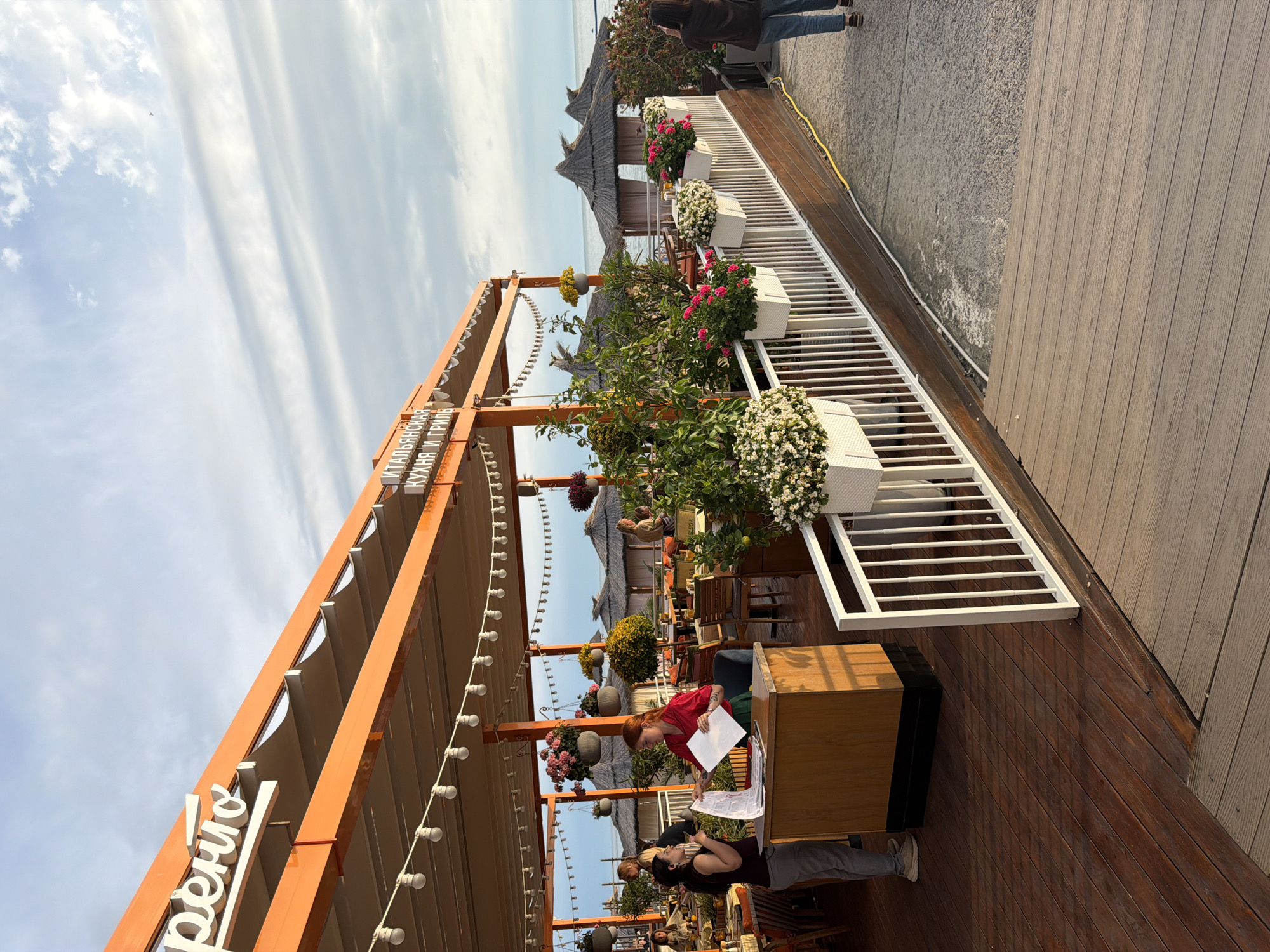 This is an outdoor scene at a restaurant, likely in Sochi, Russia, given the text on the awning. The restaurant appears to be located by the sea, with thatched-roof huts visible in the background and the sea beyond. The awning is orange and has white lettering that reads "FRENTO" and "ITALIAN CUISINE AND GRILL". There are tables and chairs set up for dining, and several people are present. Two women are standing at a wooden counter; one, in a red shirt, is holding a white sheet of paper, and the other, in a dark top, appears to be speaking to her. Several potted plants with white and pink flowers are displayed along a white railing. The ground is a wooden deck. The sky is partly cloudy.