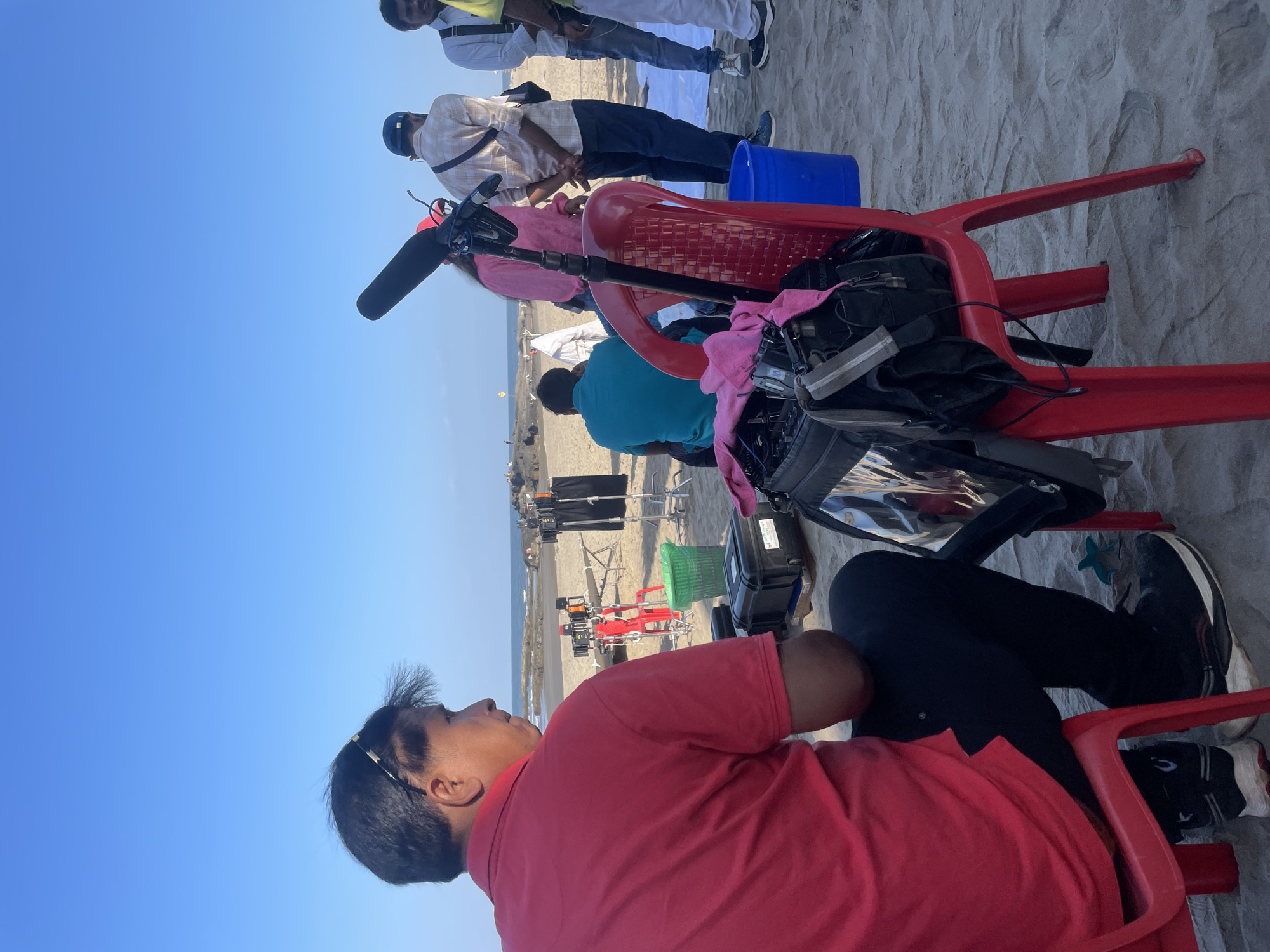 The image shows a film set on a beach in Chopdem, India. In the foreground, a man wearing a red shirt and black pants sits on a red plastic chair. Behind him, another red chair holds a sound recording device with a microphone. Further back, a group of people stand near white sheets on the sand. Film equipment, including lights and stands, are visible in the distance. The sky is clear blue.