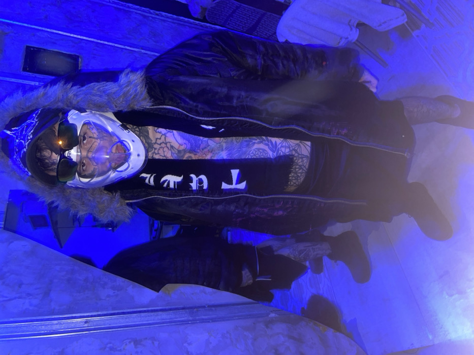 A person wearing a black jacket with a fur-trimmed hood and a white mask with sunglasses stands in a room with blue lighting. The person has visible tattoos on their chest and arms, and is wearing a black shirt with white lettering. They also have black pants and black shoes. The setting appears to be indoors.