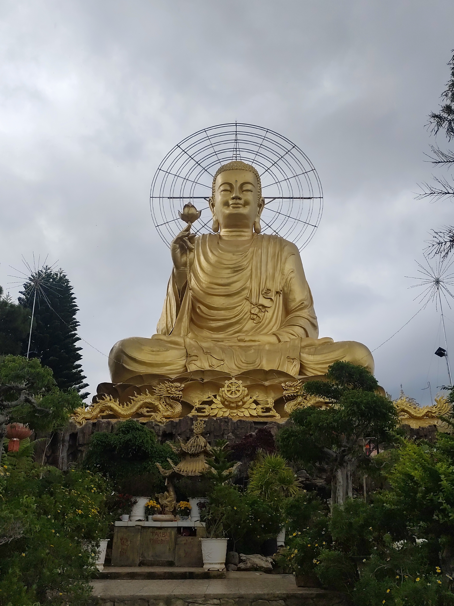 This is a full-shot, eye-level, outdoor photograph of a large, golden Buddha statue in Lâm Viên - Đà Lạt, Vietnam. The Buddha is seated cross-legged, holding a lotus flower in its right hand, and appears to be radiating light, suggested by a halo-like structure behind its head. The statue is positioned on a tiered platform adorned with golden dragon sculptures and intricate detailing. In the foreground, lush green trees and potted plants create a garden-like setting. The sky is overcast with scattered clouds, and some festive lights are visible on tall poles to the left and right of the statue. The overall ambiance is one of serenity and grandeur.