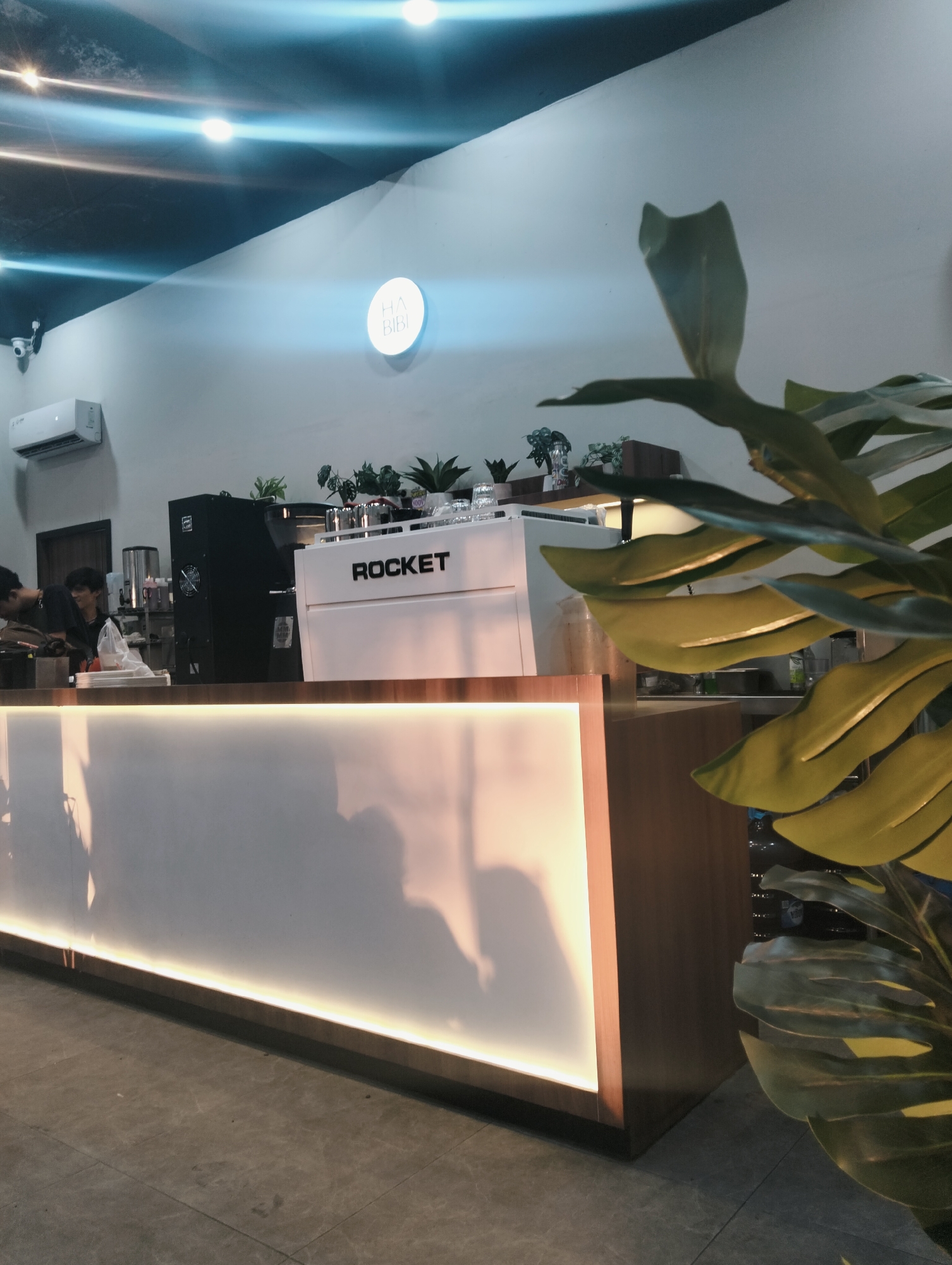 In the image, a modern cafe interior is depicted, likely located in the city of Medan, Indonesia. The scene focuses on a brightly lit counter area with a white and wood finish. A prominent white espresso machine with "ROCKET" branded on it is positioned on the counter. To the left of the espresso machine, a black commercial coffee grinder is visible, along with a stainless steel container and other cafe equipment. Behind the counter, a white circular sign with the text "HA BIBI" in blue lettering is mounted on the light-colored wall. An air conditioning unit is mounted high on the wall to the left, and a security camera is also visible in the upper left portion of the frame. In the foreground, a large, lush green plant with broad leaves partially obscures the right side of the counter. Two individuals, likely cafe staff, are partially visible behind the counter, engaged in their work. The flooring appears to be a dark grey tile. The overall ambiance suggests a clean, contemporary coffee shop.
