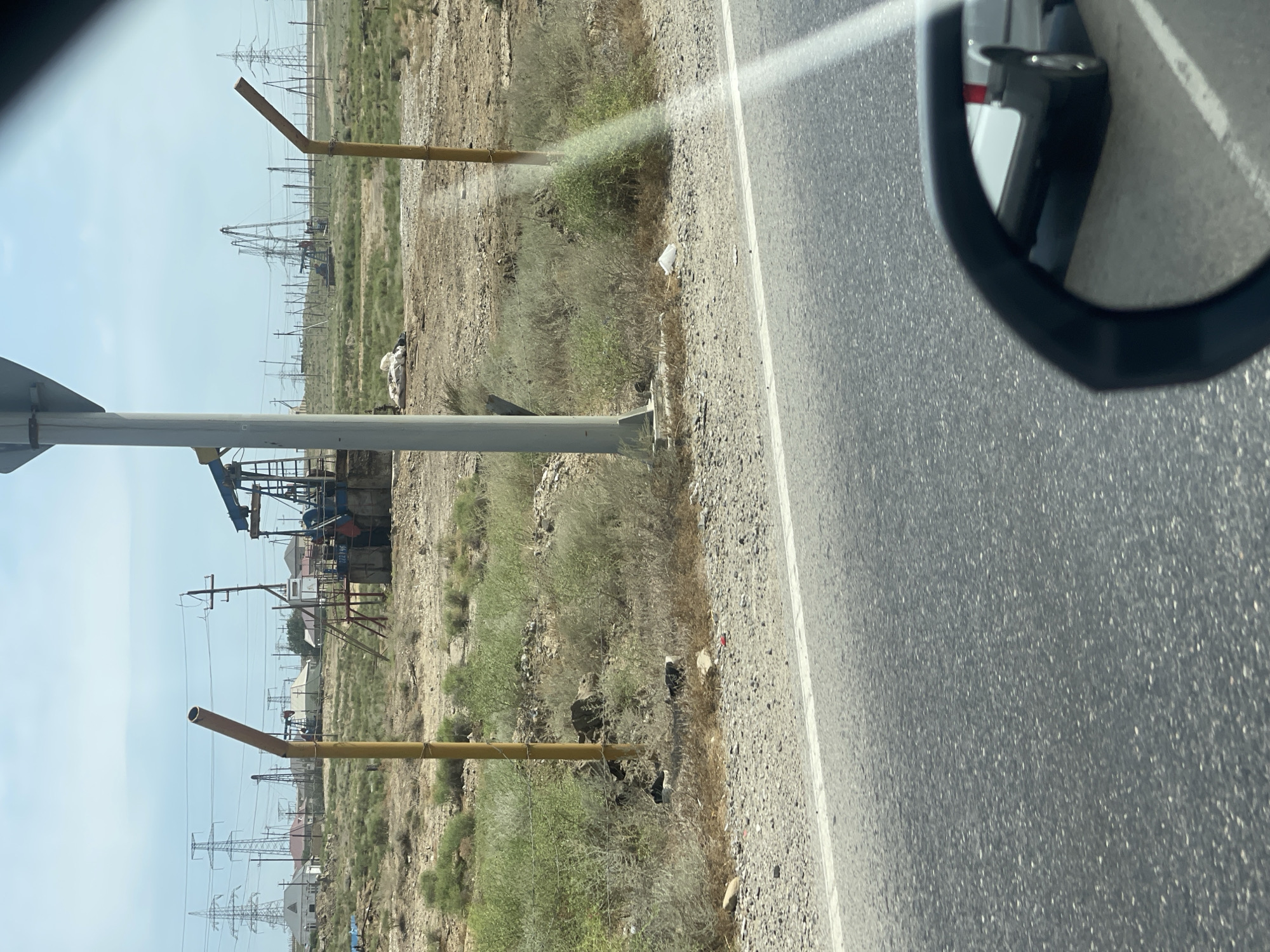 The image depicts an industrial landscape with multiple oil pumpjacks in the background, likely in the city of Digah qəsəbəsi, Azerbaijan, given the context. In the foreground, a grey asphalt road runs horizontally, with a white line marking its edge. The roadside is covered with gravel and sparse vegetation. Two yellow, angled metal poles are visible on the left and right sides of the frame, partially obscuring the view of the oilfield. A grey utility pole stands between the two yellow poles. In the middle ground, several oil pumpjacks are operational, along with other industrial equipment and power lines supported by tall metal towers. Scattered buildings, possibly residential, are visible behind the oilfield. The sky is clear and blue. The image appears to be taken from a vehicle, as a car's side-view mirror is partially visible in the lower right corner, reflecting a white vehicle. Some debris, including white plastic bags, is scattered along the roadside.