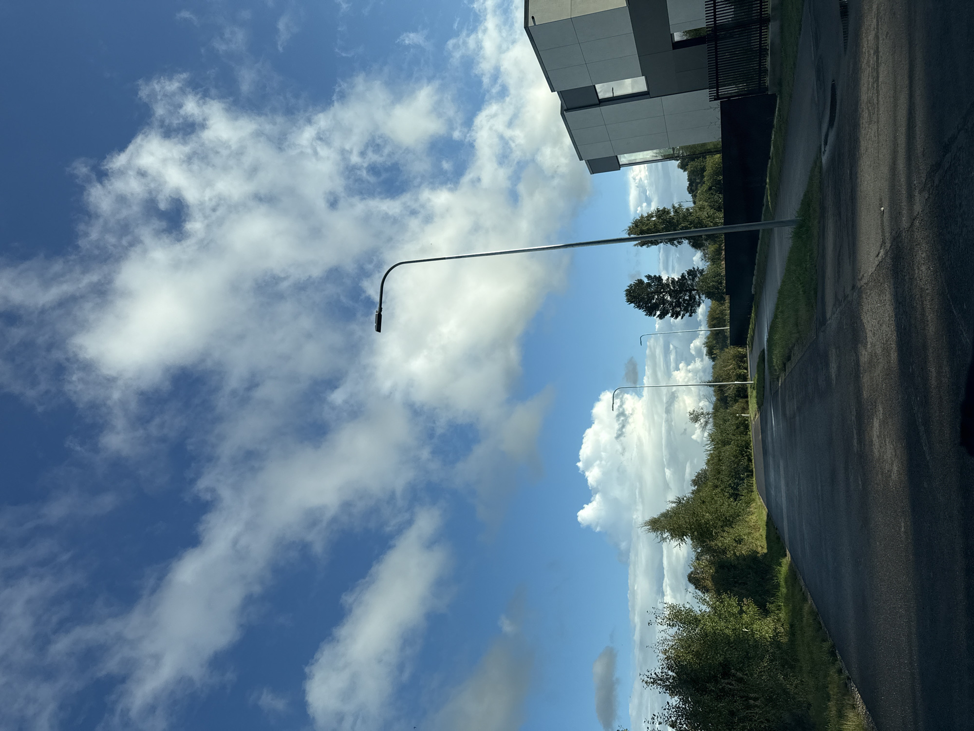 The image shows a paved road in Harkujärve, Estonia. The road is lined with trees and lush green vegetation on both sides. On the right side of the road, there is a modern building with a gray facade and large windows, and a black metal fence. A tall street lamp stands near the building. The sky is a vibrant blue, with scattered white clouds. The road appears wet in places, possibly from recent rain. The scene is devoid of people.