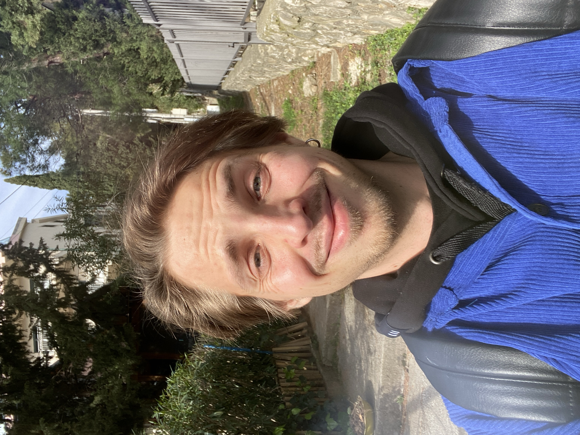A man with light brown hair, wearing a black hoodie under a blue corduroy shirt and a black leather jacket, is taking a selfie outdoors in Šušanj, Montenegro. He has an earring in his left ear. The background features a stone wall, vegetation (trees, bushes), a metal fence, and buildings in the distance. The location is sunny.