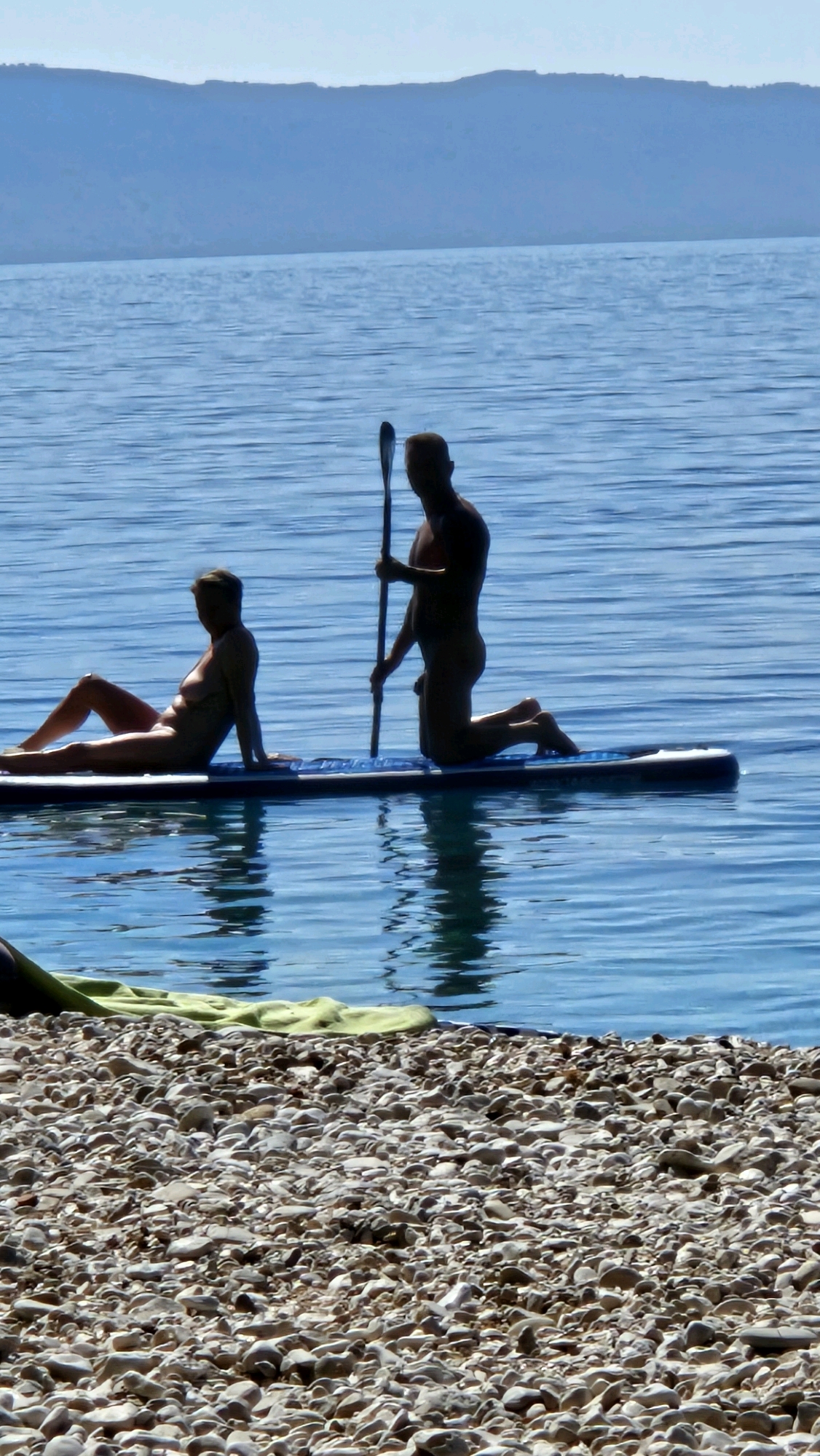 Two nude figures are on a paddleboard in the Adriatic Sea, with the coastline of Općina Podgora, Croatia, in the background. The figure on the left is a woman lying on her side, facing left. The figure on the right is a man kneeling on the paddleboard, holding a paddle. The water is blue and calm, and a pebble beach is visible in the foreground. The sky is clear and blue.