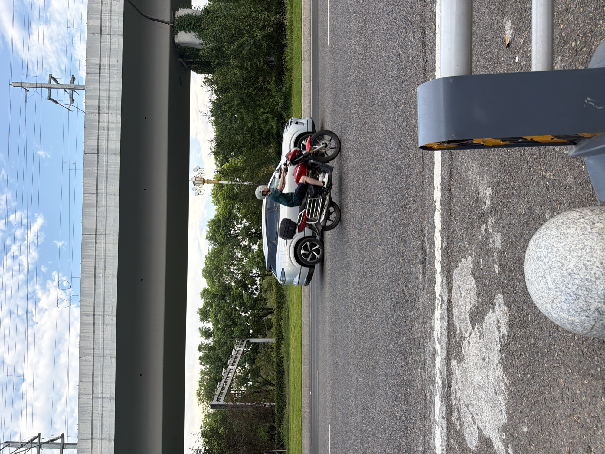 A motorcycle rider wearing a green shirt and a white helmet is riding a red motorcycle on a paved road in Jiangjin, China. The motorcycle has a black storage box on the back. To the right of the motorcycle, a silver SUV is driving in the same direction. In the background, a concrete overpass stretches across the scene, supported by pillars. Power lines are visible above the overpass. Lush green trees and bushes line the sides of the road. The sky is partly cloudy. In the foreground, a grey, speckled spherical bollard is partially visible on the left, and a grey and yellow traffic barrier is on the right. The road surface shows some signs of wear and has a white lane marking.