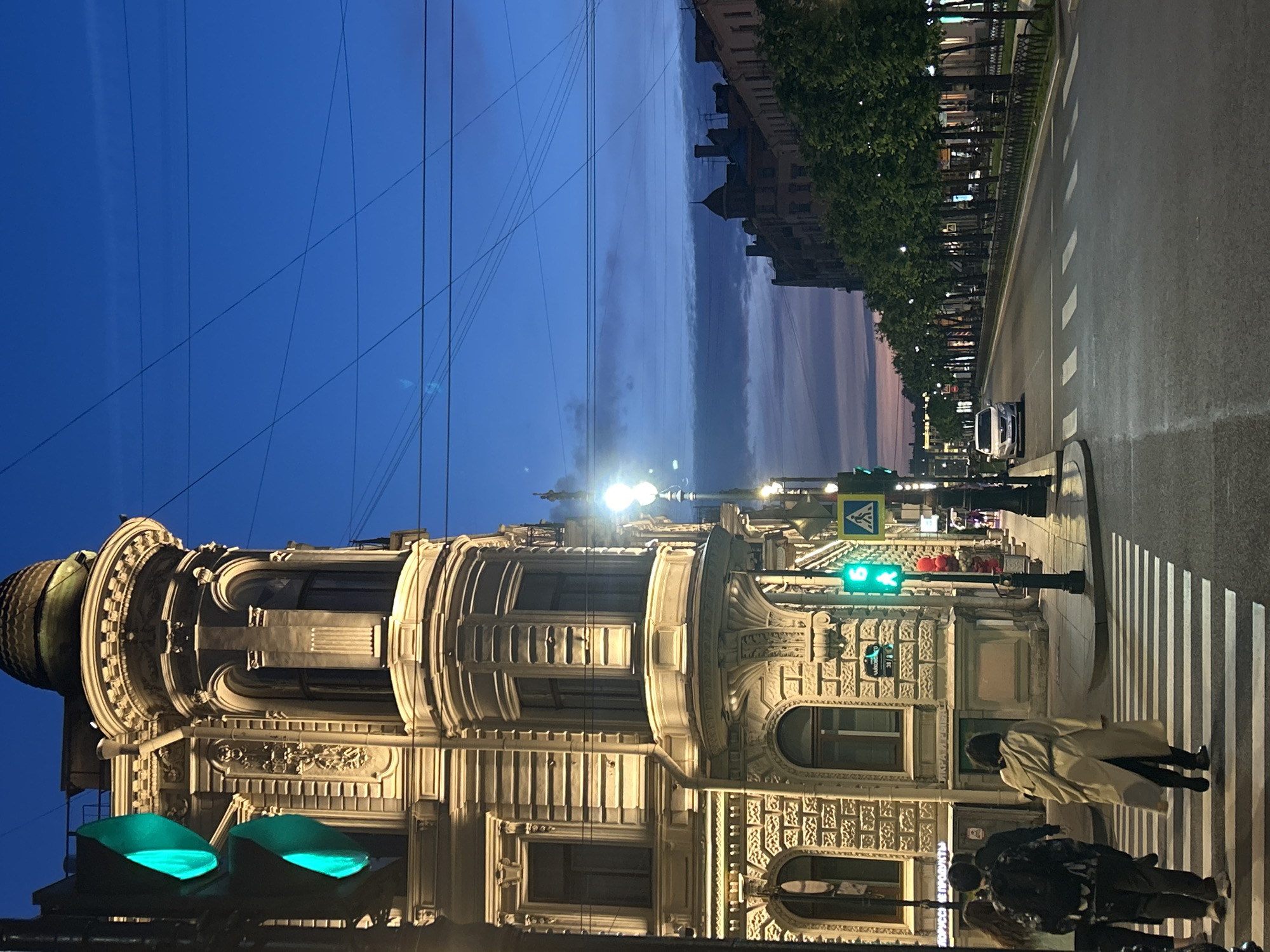 In the city of Unknown, Unknown, at dusk, an ornate building with intricate carvings and a domed roof stands on the left side of the image. A traffic light with a green signal is visible to the left of the building, illuminating the scene. Several individuals are crossing a crosswalk in front of the building, with one person in a tan trench coat walking away from the camera. A street lined with trees and a black metal fence stretches down the right side of the image, with a car parked on the side of the road. The sky is a deep blue with hints of pink and purple as the sun sets, and power lines are crisscrossed overhead.