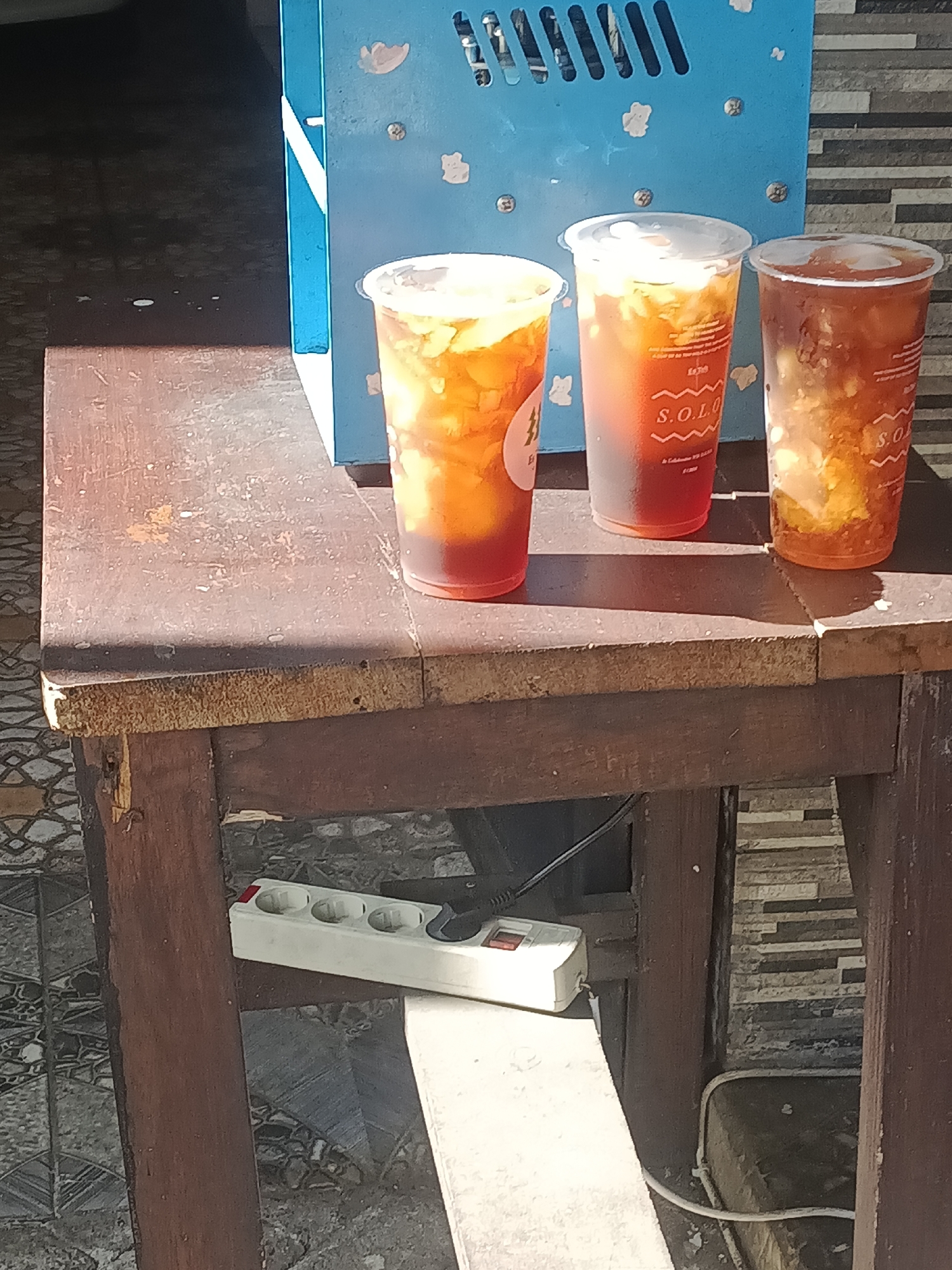 The image shows three plastic cups filled with iced tea and fruit pieces, labeled with the word "SOLO," placed on a weathered wooden table. The table sits outdoors, likely in Prigen, Indonesia, given the prompt's context. A light blue object with decorative cutouts is positioned behind the cups. Below the table's surface, an electrical power strip is visible, along with a white supporting structure. The ground surrounding the table is paved, and a wall with a horizontal pattern is in the background. There are no people visible.