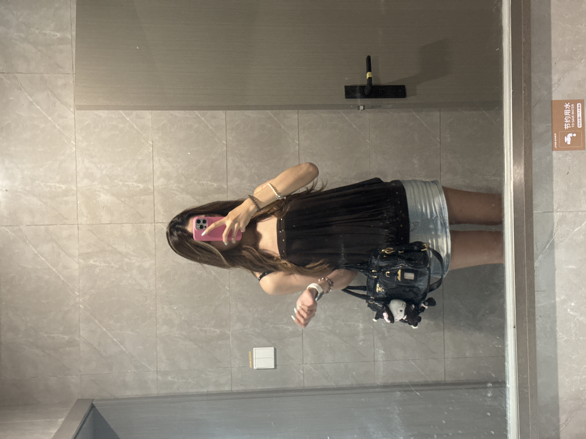 A woman is taking a selfie in a bathroom mirror. The location is in Nan'an District, China. The woman is wearing a black top and a denim skirt and carrying a black purse. She has long brown hair and is wearing bracelets on her left wrist. She is holding a pink phone in front of her face. The bathroom has gray tile walls and a toilet paper dispenser. A sign that reads "To Save Water" in English and Chinese is visible on the right side of the mirror. A squeegee is on the wall.