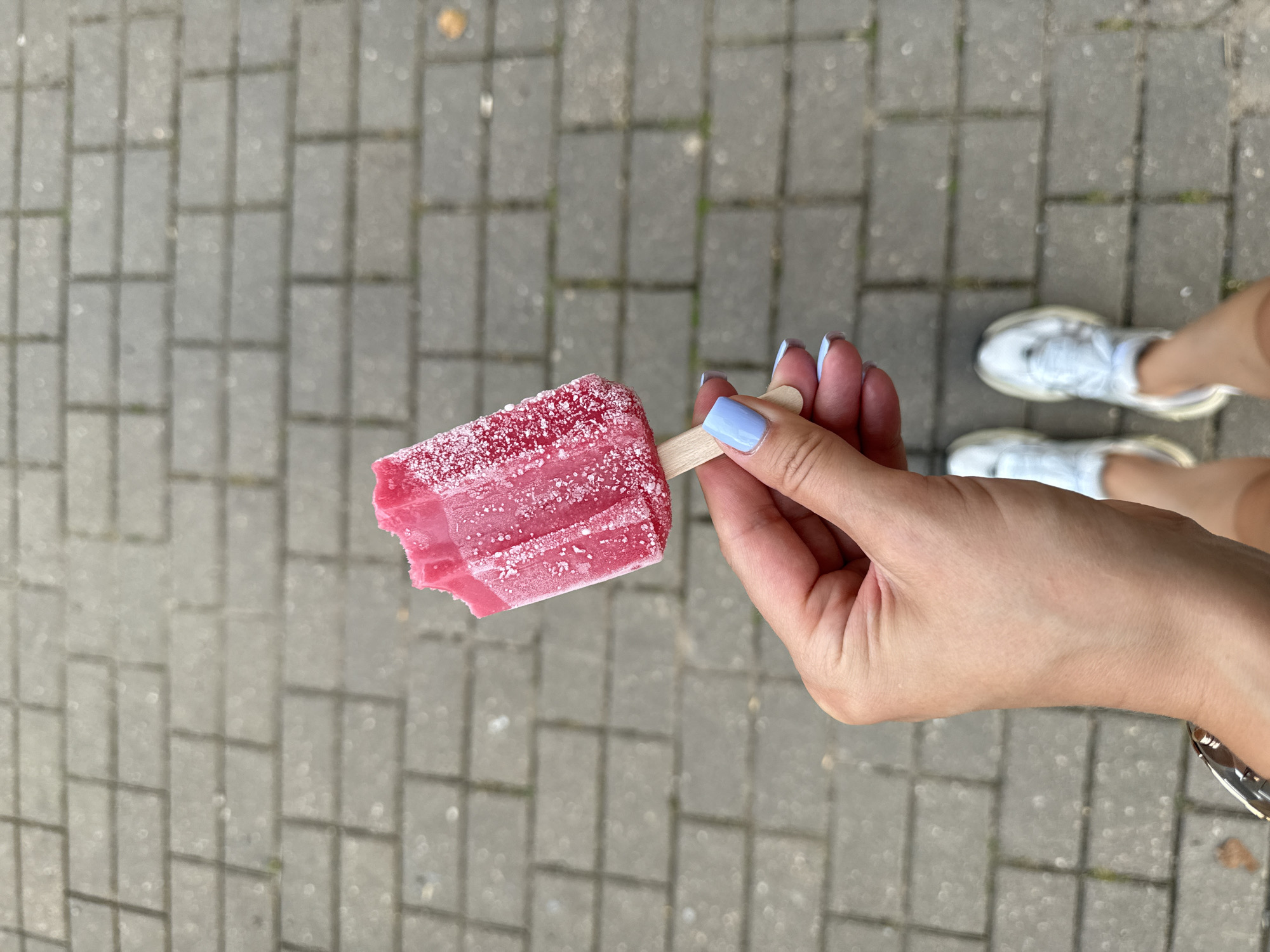 A first-person perspective shot shows a hand holding a partially eaten red popsicle with a wooden stick. The popsicle is dusted with white sugar crystals. The hand has light blue nail polish and is resting on a paved surface made of rectangular grey bricks. In the background, a person's legs are visible, wearing light blue sneakers. The scene appears to be outdoors on a sidewalk.
