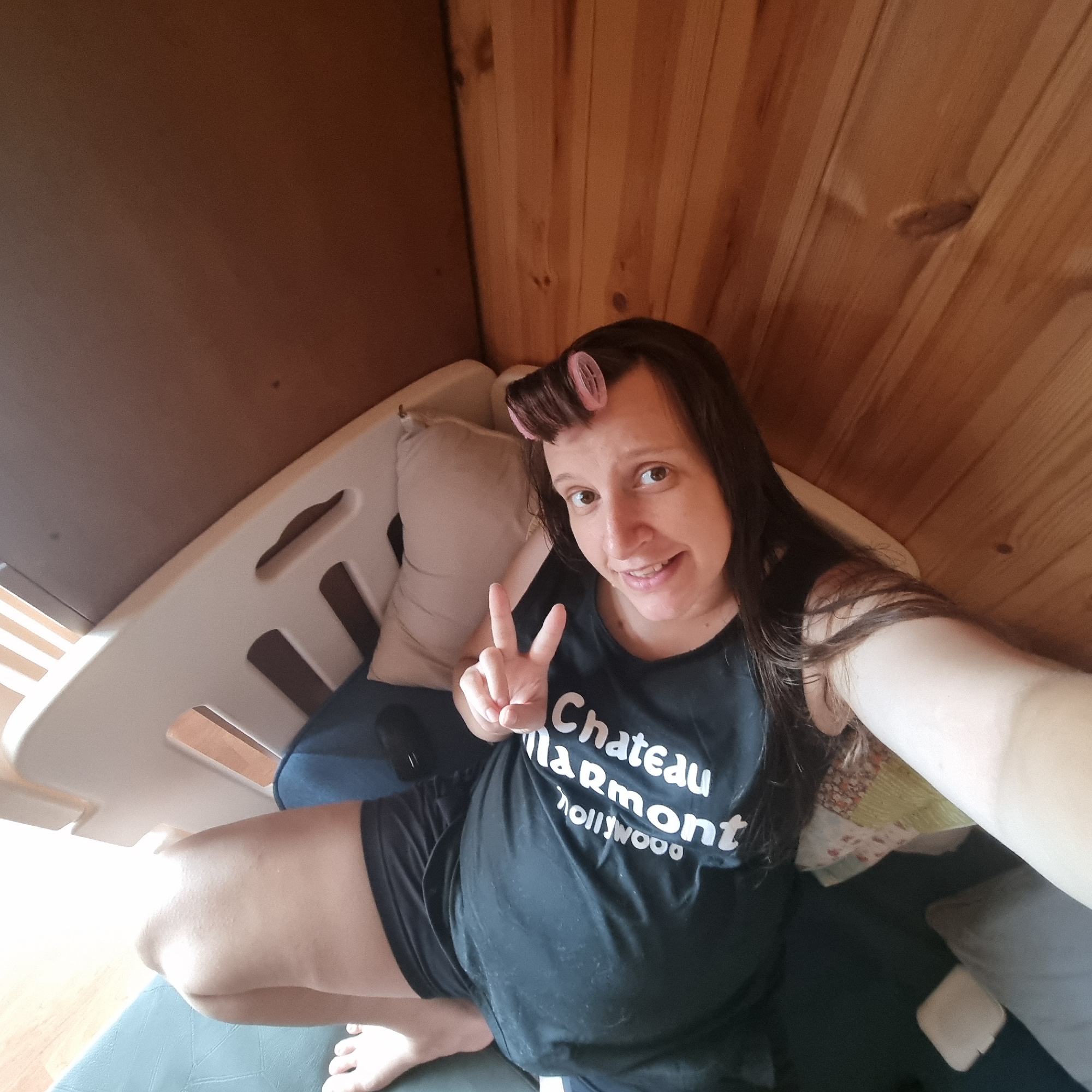 A woman is sitting in a beige plastic crib or playpen. She has pink hair rollers in her bangs. She is wearing a black tank top with "Chateau Marmont Hollywood" printed on it and black shorts. She is holding up two fingers in a peace sign. The background consists of wooden paneling and a darker brown wall. The crib is against the wooden paneling. She is smiling and looking at the camera.