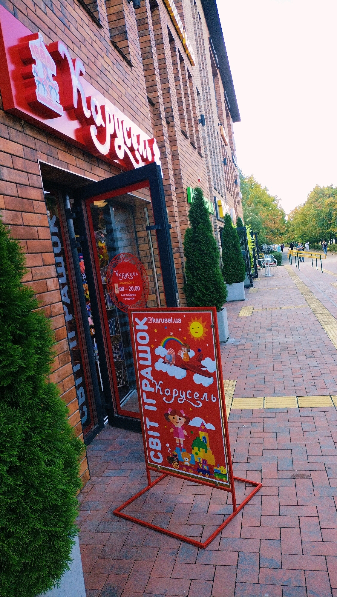 The image captures the exterior of a toy store named "Карусель" (Carousel) located in Horishni Plavni, Ukraine. A red sign with white lettering advertising "СВІТ ІГРАШОК" (World of Toys) and "@karusel.ua" is prominently displayed on the sidewalk in front of the store. The sign features a colorful illustration of children on a flying airplane amidst clouds and a rainbow, with a happy sun above. The store's facade is made of brick, and a large red sign with "Карусель" in white script is visible above the entrance, which is partially open. Inside the store, shelves stocked with toys are visible through the glass. Two evergreen trees flank the entrance. The sidewalk is paved with red and grey bricks, and yellow tactile paving is present. In the background, people can be seen walking along the sidewalk and trees with autumn foliage are visible. The sky is overcast.