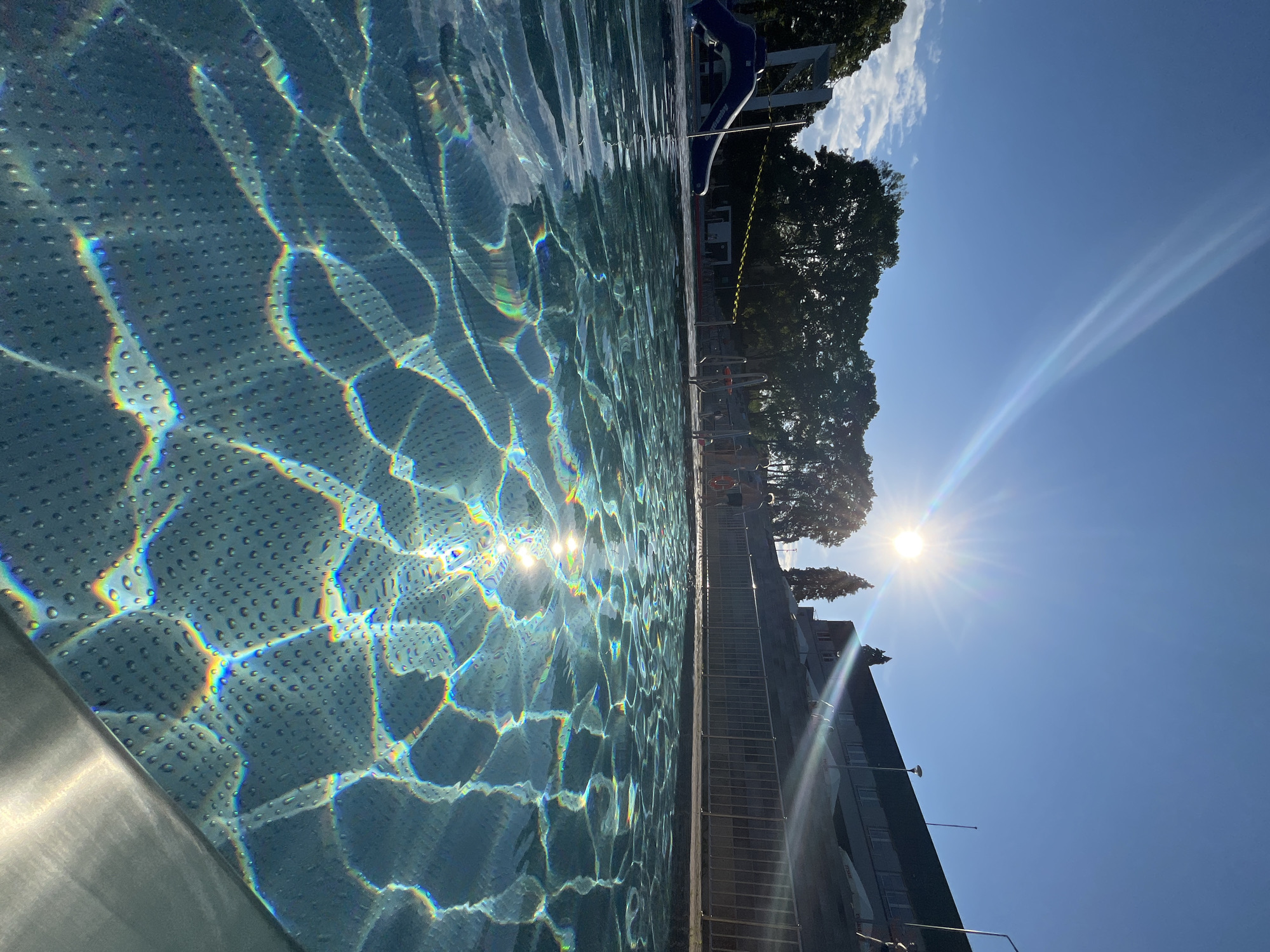 The image depicts an outdoor swimming pool scene on a sunny day in the city of Unknown, Unknown. The perspective is low, looking across the pool water towards the horizon.
The sky in the upper half of the image is clear and bright blue, with the sun prominently visible in the upper-right quadrant, emitting strong sunburst rays and lens flare that extend across the sky. A few small, white cumulus clouds are present in the upper-left.
In the mid-ground, a line of dark green deciduous trees occupies the left and center, appearing as silhouettes against the bright sky. To the far left, a blue and white inflatable water slide structure is partially visible behind yellow caution tape. In the mid-right, a multi-story building with a dark roof and multiple windows is visible, partially obscured by the sun's glare and some slender trees (possibly cypresses).
Several individuals are present in the mid-ground, appearing as dark silhouettes due to the backlighting. They are positioned near a light-colored metal fence that runs parallel to the pool edge, separating the pool area from an elevated section where the building stands. One person near the right side of the fence appears to be holding a red lifebuoy. Their exact actions are indistinct.
The foreground is dominated by the clear, blue water of the swimming pool. The surface of the water shows ripples and waves, off which the strong sunlight reflects intensely. These reflections create numerous fragmented, iridescent, rainbow-colored patterns of light that shimmer across the textured pool bottom. The pool bottom itself has a distinct pattern of small, raised bumps or dots, possibly for anti-slip purposes. The lower-right corner shows a metallic, possibly chrome, edge of the pool, likely a coping or gutter.
