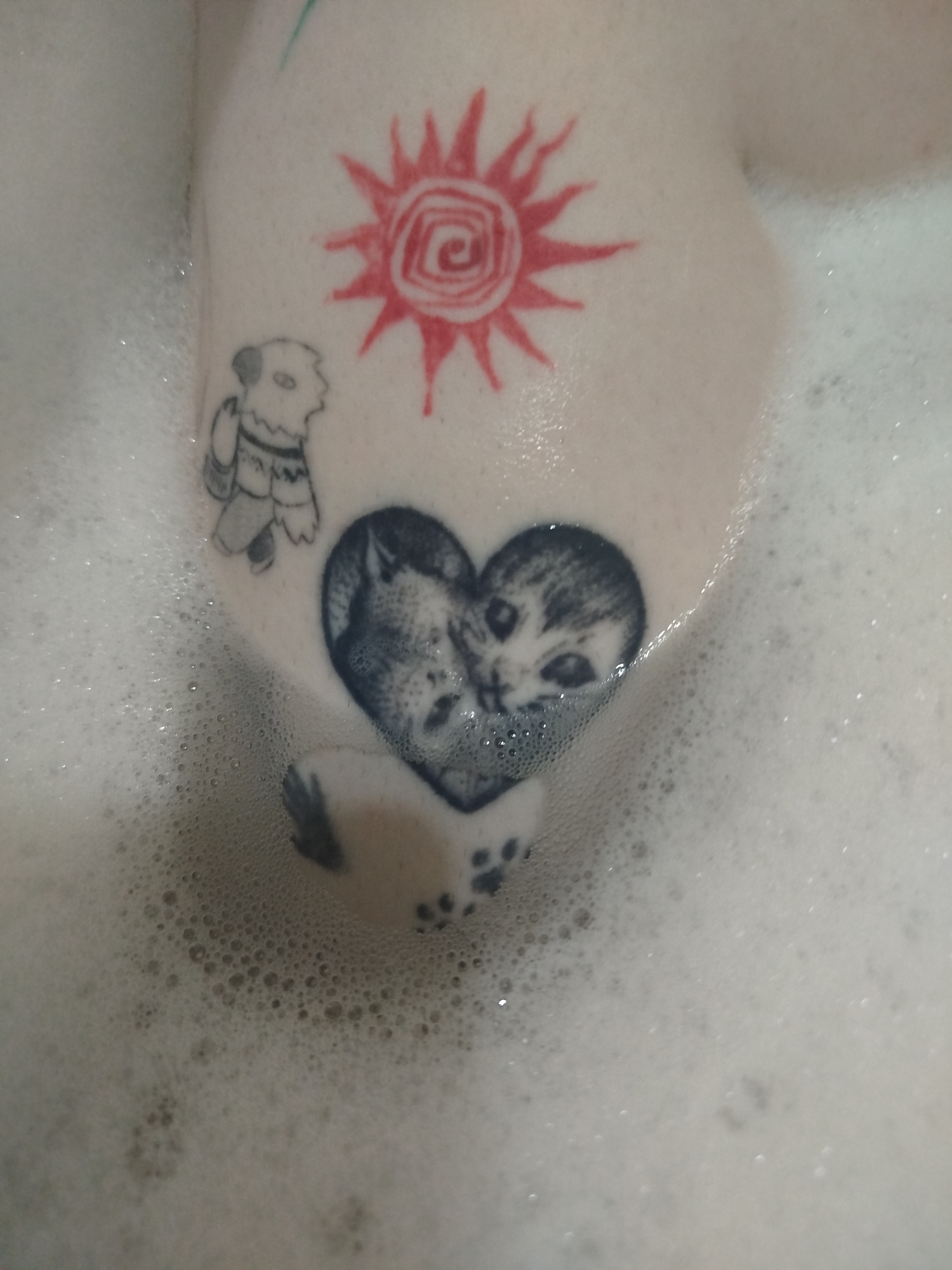 The image shows a body part submerged in a tub filled with bubbly water. Several tattoos are visible on the skin. There is a red tattoo of a sun, a black and white tattoo of a character or creature with a sweater, and a black and white tattoo of two cats forming a heart shape. One paw is also visible on the body part. The location appears to be a bathroom, as evidenced by the bathtub and bubbly water. The city and state are unknown.