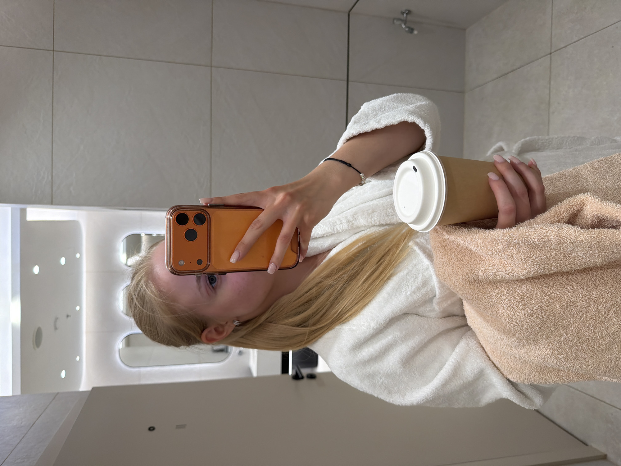 A female stands in a bathroom in Unknown, Unknown. She holds a brown smartphone in her left hand to take a selfie. She is wearing a white bathrobe, and in her right hand, she holds a brown coffee cup with a white lid. A tan-colored towel is draped over her right shoulder. The bathroom has light gray tile walls and a chrome showerhead in the background. A vanity with a mirror and lights is visible on the left.