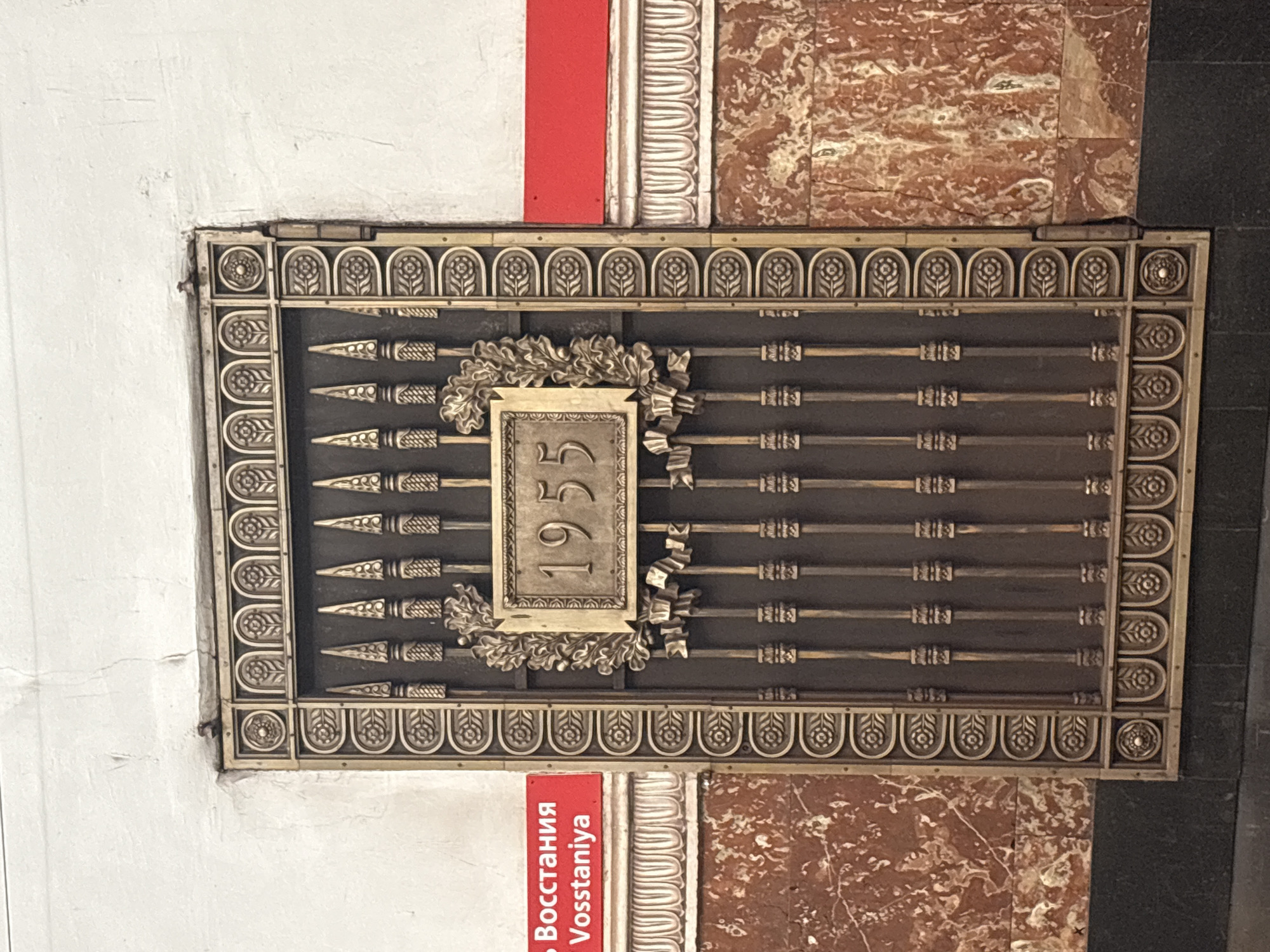 The image shows a decorative metal gate with the year "1955" inscribed on a plaque above a laurel wreath. The gate is located in the metro station "Vosstaniya" in Saint Petersburg, Russia. The station's walls are decorated with marble and red and white stripes. The gate is ornate, featuring floral motifs and spear-like elements. The plaque with the year is in a golden rectangular frame, and the numbers are embossed. The overall impression is one of historical significance and architectural grandeur.