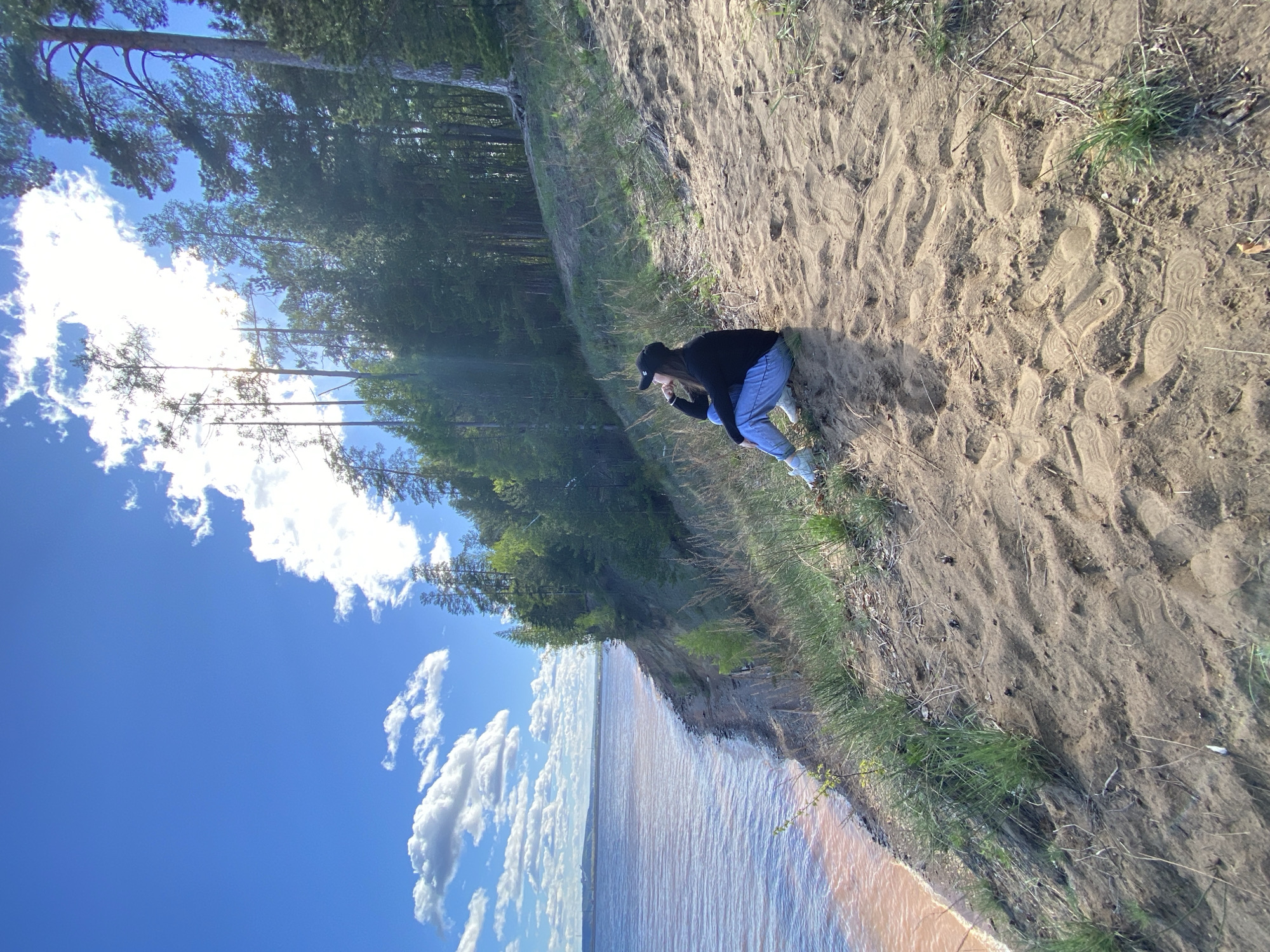 The image shows a view of a shoreline with a person on a sandy bluff, likely in Noviy, Russia. The individual, wearing a dark long-sleeved shirt, blue jeans, sneakers, and a dark baseball cap, is kneeling on the sandy slope, appearing to be looking down. The ground is sandy with scattered tufts of grass. The bluff edge overlooks water, possibly a lake or river, with a sandy-colored shoreline. Beyond the bluff, a dense forest is visible. The sky is bright blue with scattered cumulus clouds. The image is slightly rotated counterclockwise.