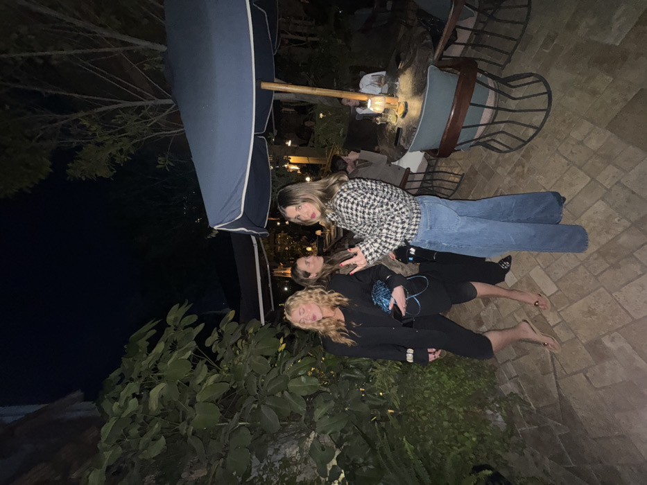 Three young women are laying on a stone patio under a large blue umbrella at night. The woman on top wears a black and white houndstooth jacket and jeans. The woman in the middle wears all black, and the woman on the bottom has blonde hair and wears all black with bare feet. Green plants are to the left, and chairs and other people are visible in the background.