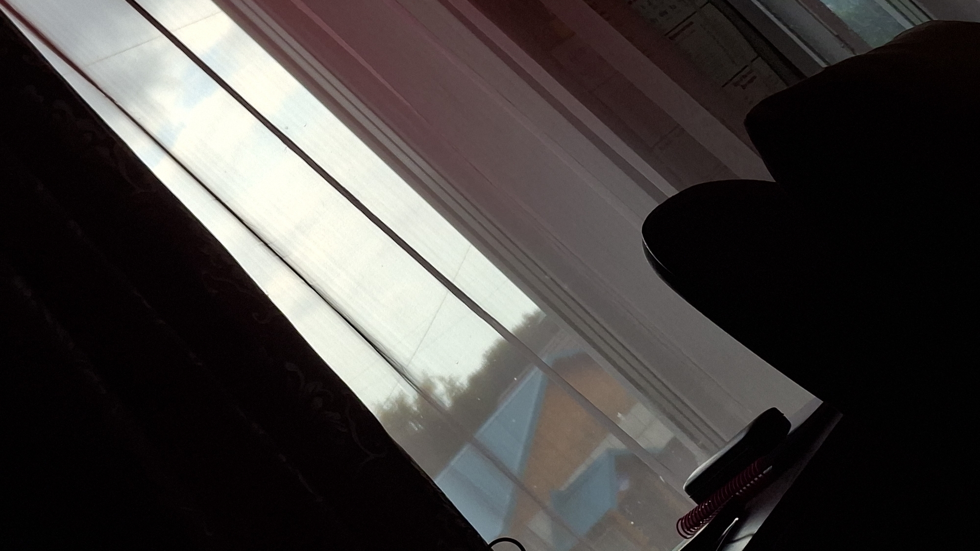 A dimly lit interior scene is captured from a low angle, looking towards a window covered by sheer, vertically striped curtains. Sunlight streams through the translucent fabric, creating bright, elongated rectangles of light and casting shadows from the room's elements.
In the foreground, to the left, a dark, heavy curtain is visible, obscuring much of the room and creating a strong contrast with the illuminated window. To the right, the dark silhouette of a chair or sofa is prominent, its form softened by the low light. Resting on a surface in the lower right corner of the frame is a notebook with a red spiral binding, partially obscured by the darkness.
Through the sheer curtains, the exterior environment is faintly visible. Hints of trees and the roofs of buildings with a blue and orange color scheme suggest a residential area. The quality of light, with its soft diffusion and long shadows, indicates it is likely late afternoon or early evening. The overall mood is quiet and contemplative, with the play of light and shadow creating a sense of depth and mystery. No text is visible in the image. The setting appears to be indoors, possibly a living room or study, with a view of the outside world.