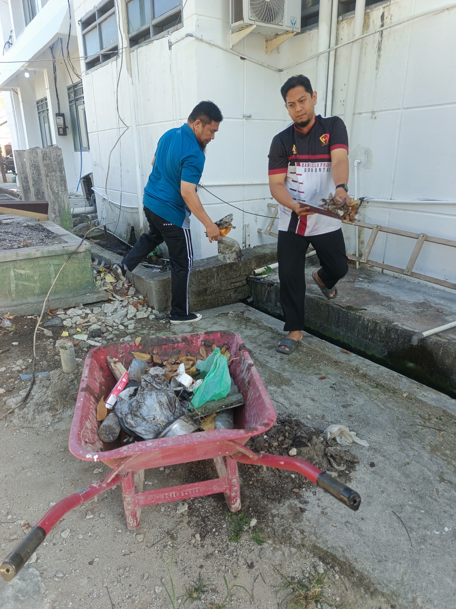 Two men are engaged in a cleanup operation outdoors. The man on the left, wearing a blue shirt and black pants with white stripes, is using a tool to gather debris. The man on the right, in a black and white shirt with red accents that reads "BAWASLU PROVINSI GORONTALO," is holding a broom and dustpan, also collecting rubbish. In the foreground, a red wheelbarrow is filled with collected trash, including plastic bags, leaves, and other refuse. The background features a white building with visible windows and an air conditioning unit, suggesting an institutional or commercial setting. The ground is paved with concrete and gravel, with some areas showing signs of disrepair or neglect, such as broken concrete pieces scattered around. A wooden ladder is propped against the building in the background. The scene depicts a public area in Gorontalo, Indonesia, where routine maintenance or waste removal is taking place.