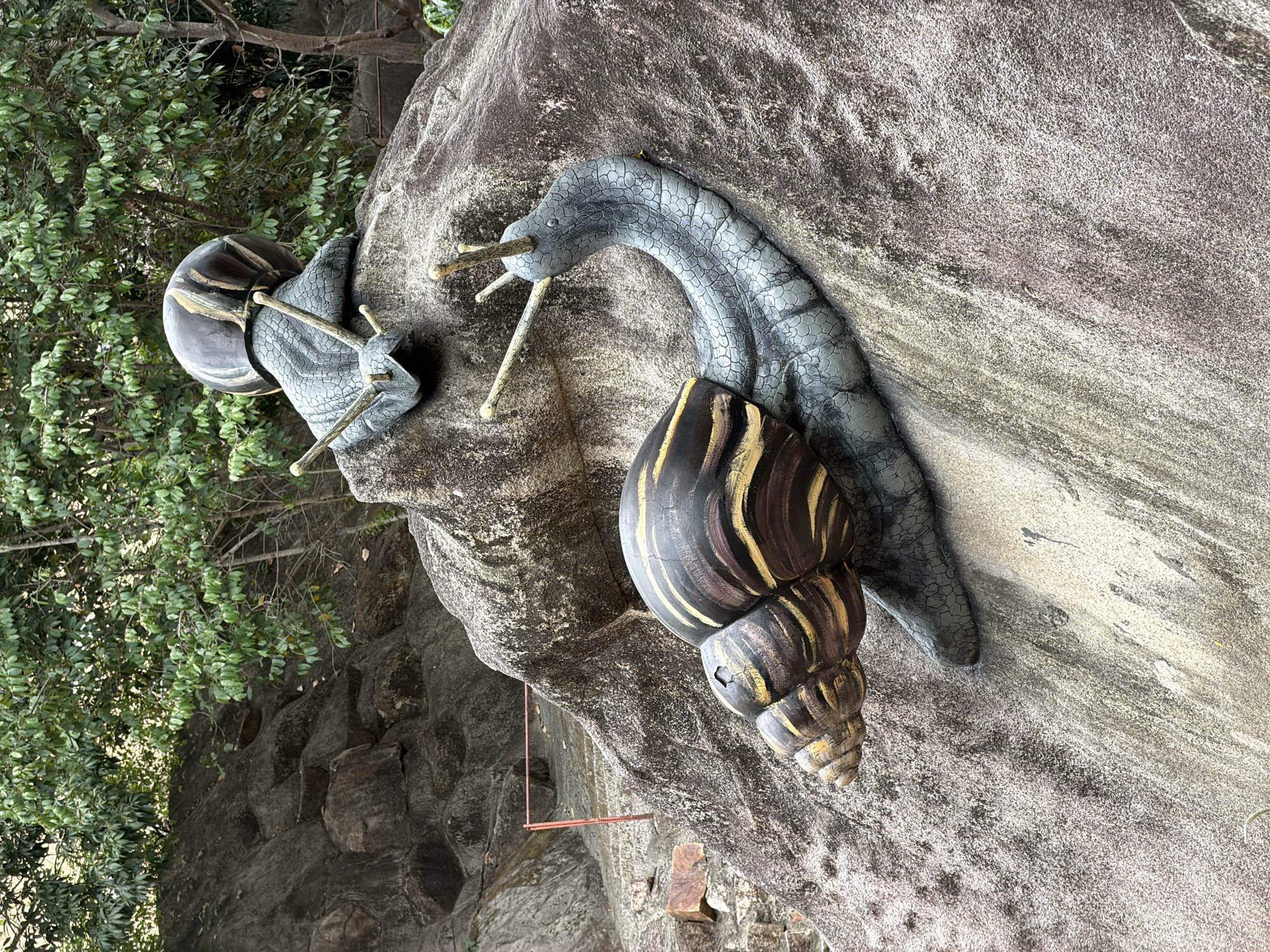 Two large, realistic snail sculptures are positioned on a rock formation. The foreground snail is larger and facing left, with its head extended and antennae raised. Its shell has alternating bands of dark brown and yellow. The snail in the background is smaller and positioned higher up on the rock, facing right, also with its antennae raised and head extended.
These sculptures are located at the entrance to the Long Son Pagoda in Nha Trang, Vietnam. The environment consists of lush green trees and foliage in the background, with a rocky, mountainous terrain surrounding the sculptures. The rock formation has an uneven, textured surface. A red rope barrier is visible in the mid-ground to the left, suggesting a pathway or boundary.