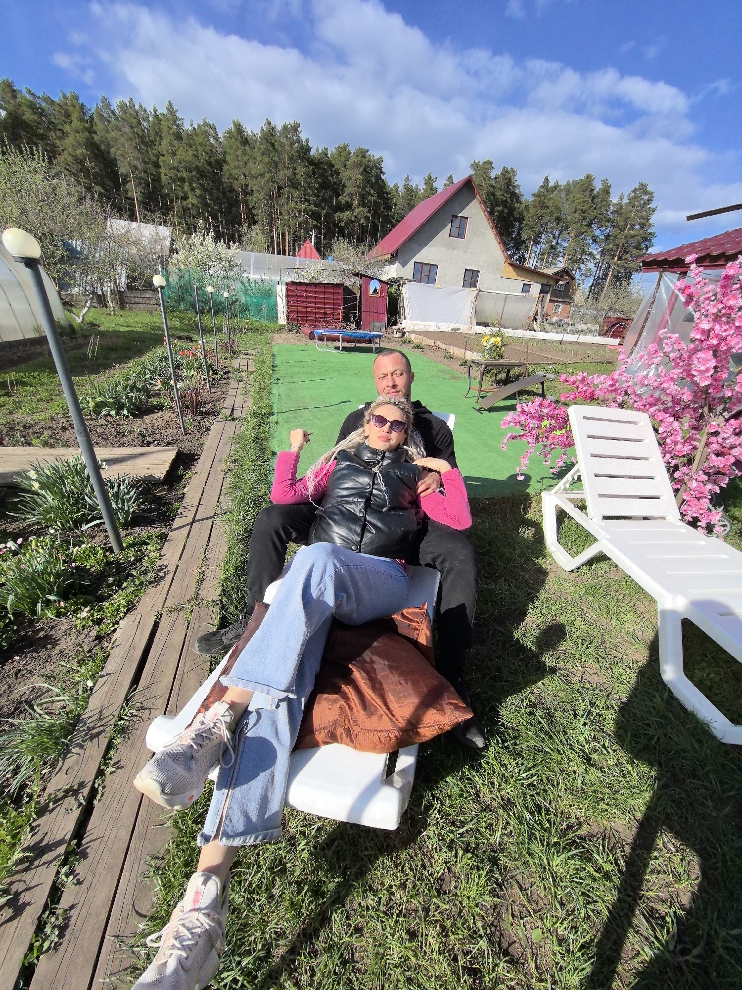 The image shows a man and a woman sitting together on a white plastic lounge chair in a garden setting. The man is wearing black clothing, while the woman has on a pink top, a black vest, and blue jeans. The woman is wearing sunglasses. The lounge chair is on the grass next to a wooden walkway. In the background, there is a house with a red roof, as well as various plants and trees. The sky is blue with scattered clouds. Given the location in the prompt, the scene is in the city of Noviy, Russia.