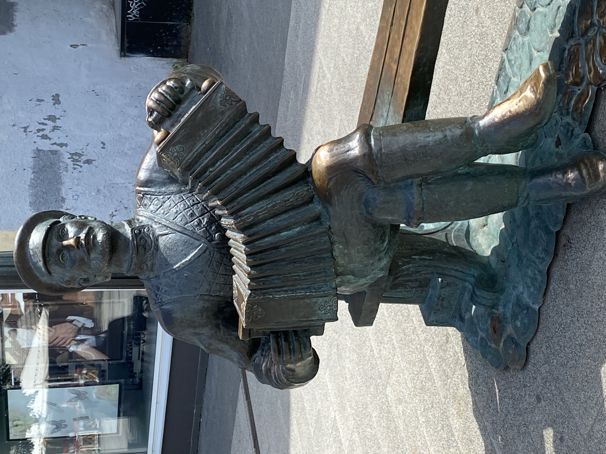 This media file features a bronze sculpture of a man playing an accordion. The sculpture is positioned outdoors, appearing to be a public installation. The man is depicted from the waist up, seated, with his legs extended. He wears a hat and a vest over a shirt, and his hands are engaged in playing the instrument. The accordion itself is detailed, with a bellows and keys visible.
The sculpture is attached to a base that seems to be made of the same material, possibly designed to resemble water or waves. Part of a wooden bench is visible behind the sculpture, suggesting it's placed in a park or public seating area. The background consists of a textured wall, possibly brick or concrete, with some graffiti visible at the top. The ground appears to be paved with light-colored, speckled stone.
The lighting suggests it is daytime, with a clear sky and strong shadows cast by the sculpture and the bench, indicating the sun is high. The overall mood conveyed by the sculpture is one of cheerful musicality and public art. There is no visible text within the image that provides information about the sculpture's title, artist, or location. The scene appears to be calm and static, capturing a moment in time of artistic expression.