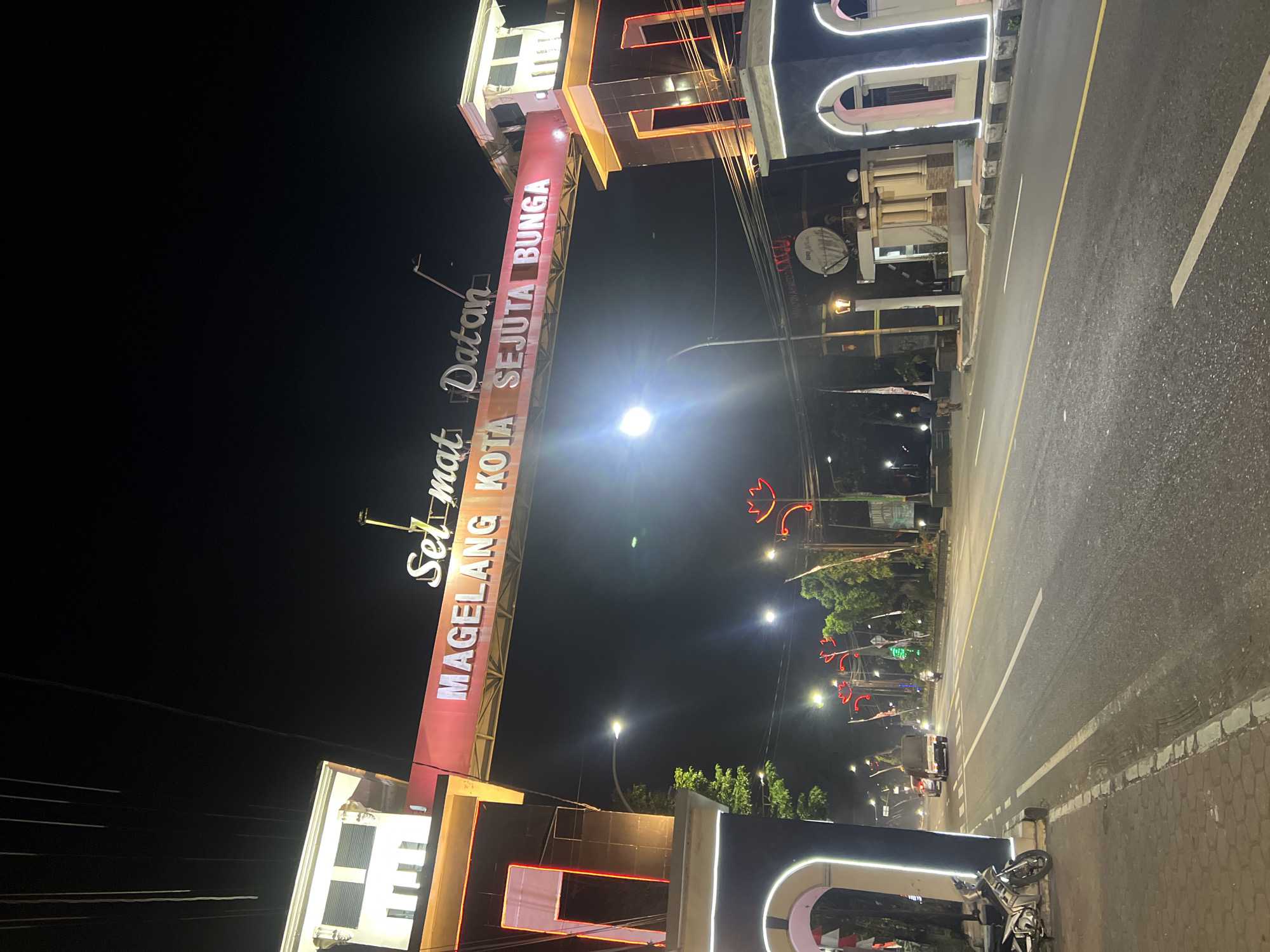 The image shows a street scene at night in Jambewangi, Indonesia, featuring a prominent archway with illuminated text that reads "SELAMAT DATANG KOTA SEJUTA BUNGA" (Welcome City of a Million Flowers). The archway is flanked by decorative pillars and is lit with red and white lights. The street itself is paved and marked with white and yellow lines. Several vehicles are visible on the road, including cars, a truck, and motorcycles. On the sidewalk to the left, a parked motorcycle is visible. Trees and streetlights line the road, and some buildings are present in the background. The overall atmosphere is illuminated and bustling with nighttime activity.