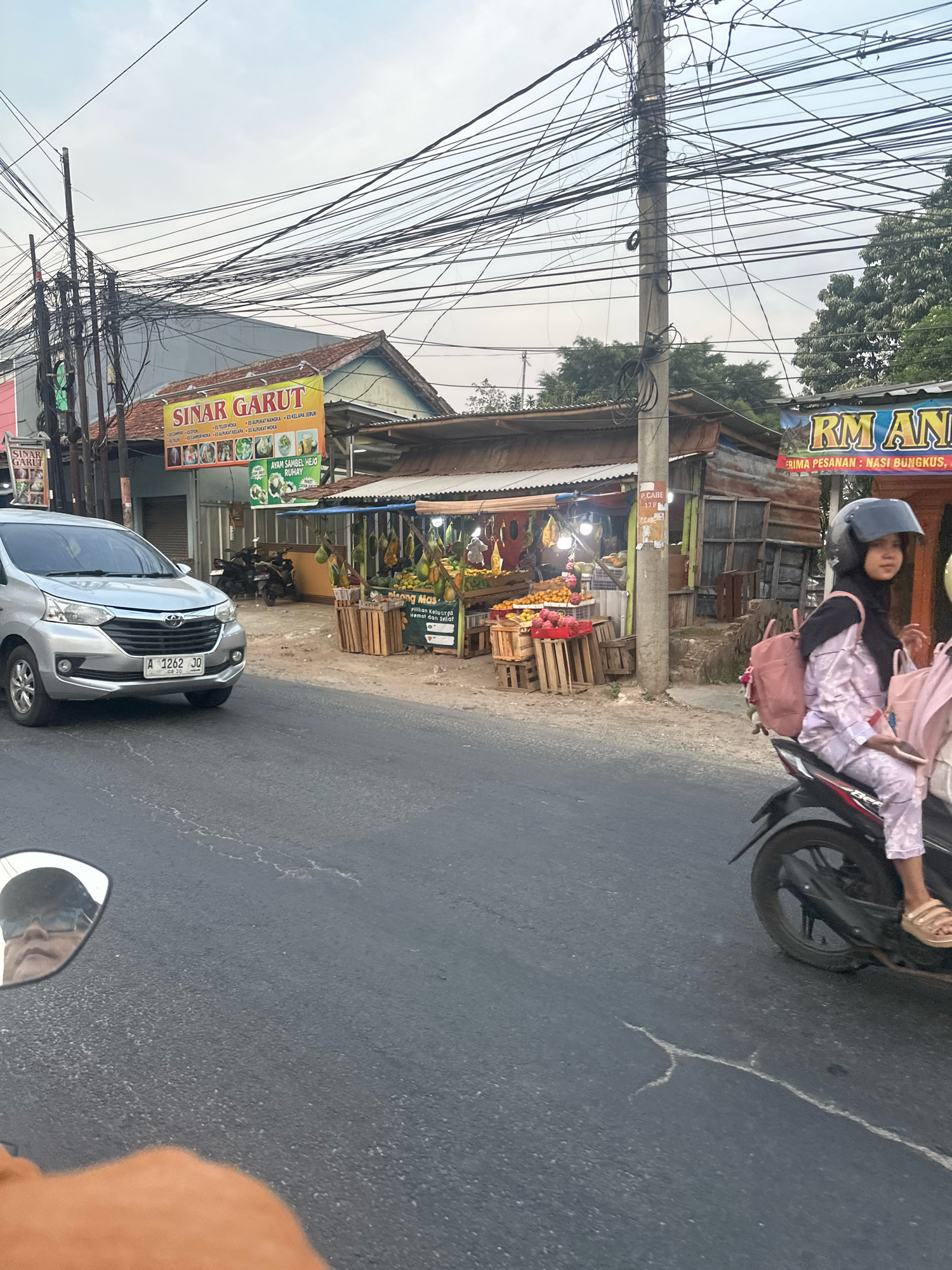 The image depicts a street scene in Cilaja, Indonesia. A silver car with license plate number A 1262 JO is parked on the left side of the road. Across the street, a fruit stall displays various fruits, including oranges and dragon fruit, in wooden crates. A woman wearing a hijab and a pink patterned outfit rides a motorcycle on the right side of the road, her face turned to the right. Overhead, a complex network of electrical wires crisscrosses the sky, suspended from utility poles. Several motorcycles are parked behind the fruit stall. A sign above the fruit stall reads "SINAR GARUT" and another sign to the right says "RM AN". The sky is overcast, suggesting a cloudy day. The overall impression is a typical street view in a bustling Indonesian town.