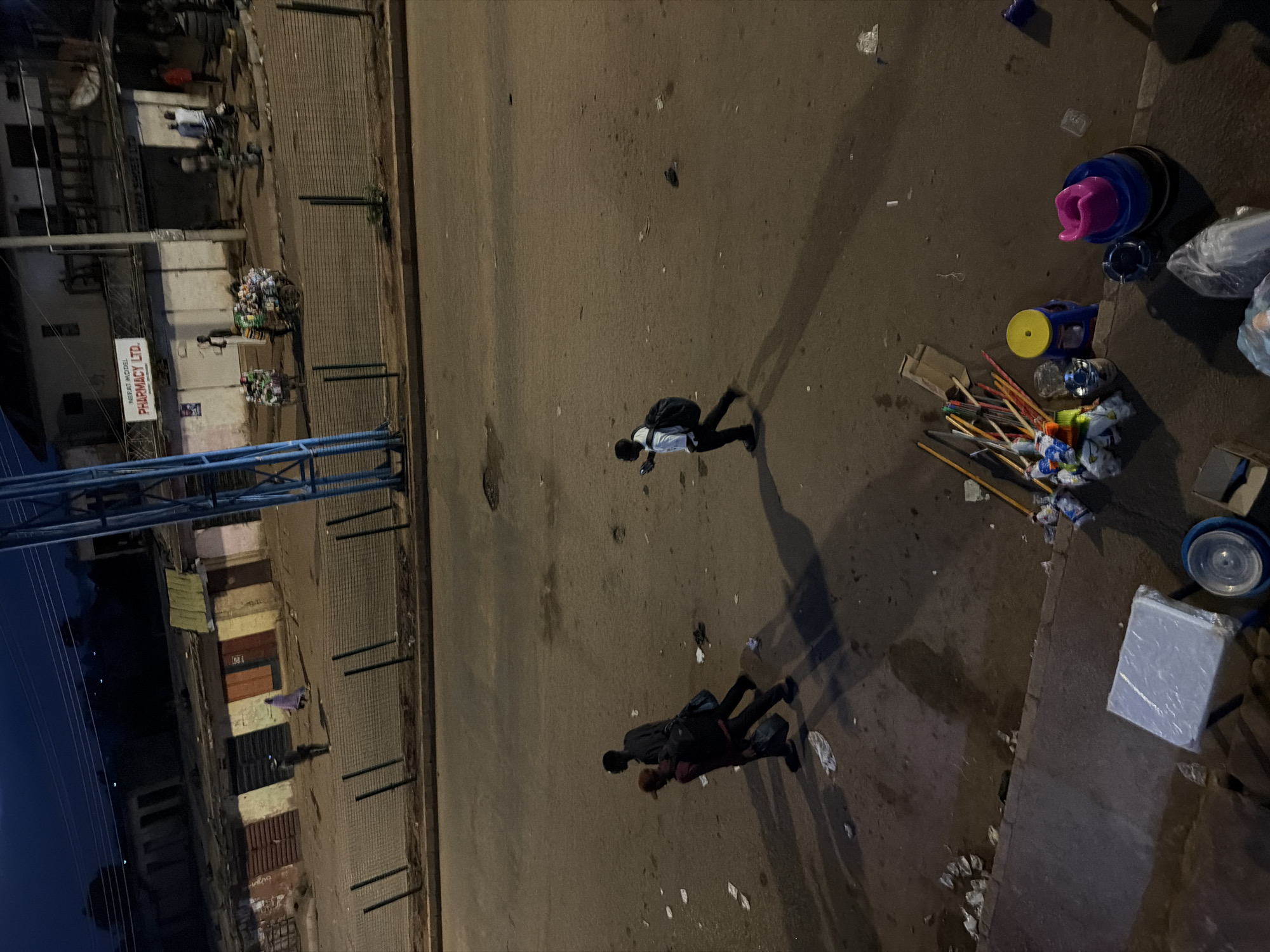 The image captures a street scene in Bukuru, Nigeria, during what appears to be nighttime or early morning due to the artificial lighting and deep shadows. The perspective is from above, looking down onto a paved street and sidewalk.
Several people are visible, walking or standing. One person, wearing a white shirt and dark pants with a backpack, is walking across the street. Further down, two people appear to be walking together, also with backpacks. The scene is somewhat cluttered, with various objects scattered on the ground and sidewalk, including what look like cleaning supplies (brooms, mops), plastic containers, and trash. There is also a stack of items wrapped in plastic, possibly merchandise.
On the left side of the image, buildings line the street. One building has a sign that reads "NBIA MODEL PHARMACY LTD." The buildings have storefronts, some of which appear to be closed. There is also a structure with blue metal bars, potentially part of a fence or canopy. The overall impression is of a commercial area at rest, possibly after business hours or in the early morning before activity begins. The lighting suggests it's not full daylight, with the shadows indicating a low light source. The atmosphere is one of quiet movement and urban life.