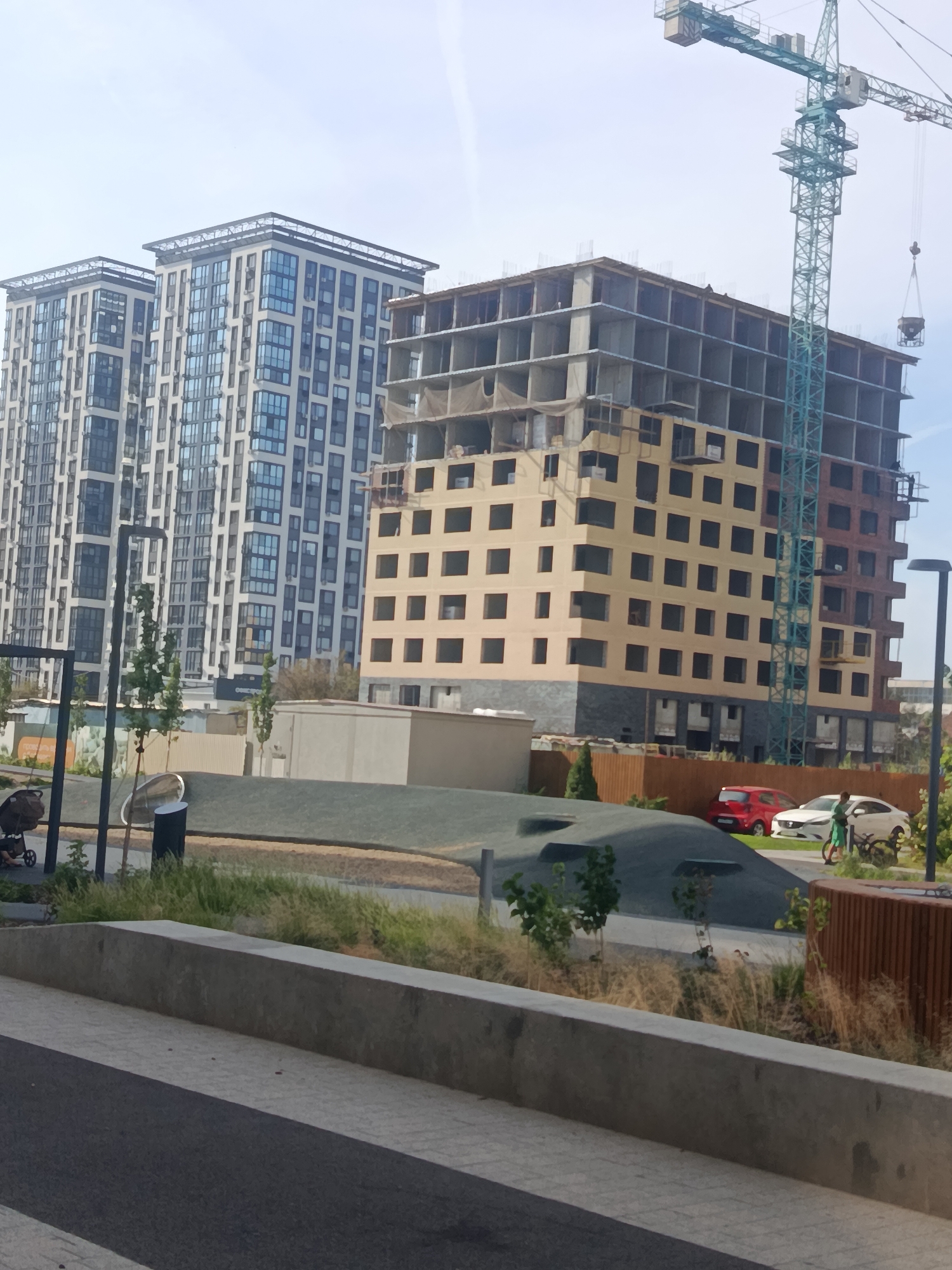 This is an outdoor medium shot photograph of a construction site in Astrakhan, Russia, featuring two completed high-rise residential buildings and a third building under construction. A large teal construction crane is visible, its jib extending over the unfinished structure. The buildings have modern architectural designs with many windows. In the foreground, there is a pathway and some landscaping, including grass and small trees. Several cars are parked near the construction site, and a person is walking on the grass next to them. The sky is clear and overcast.