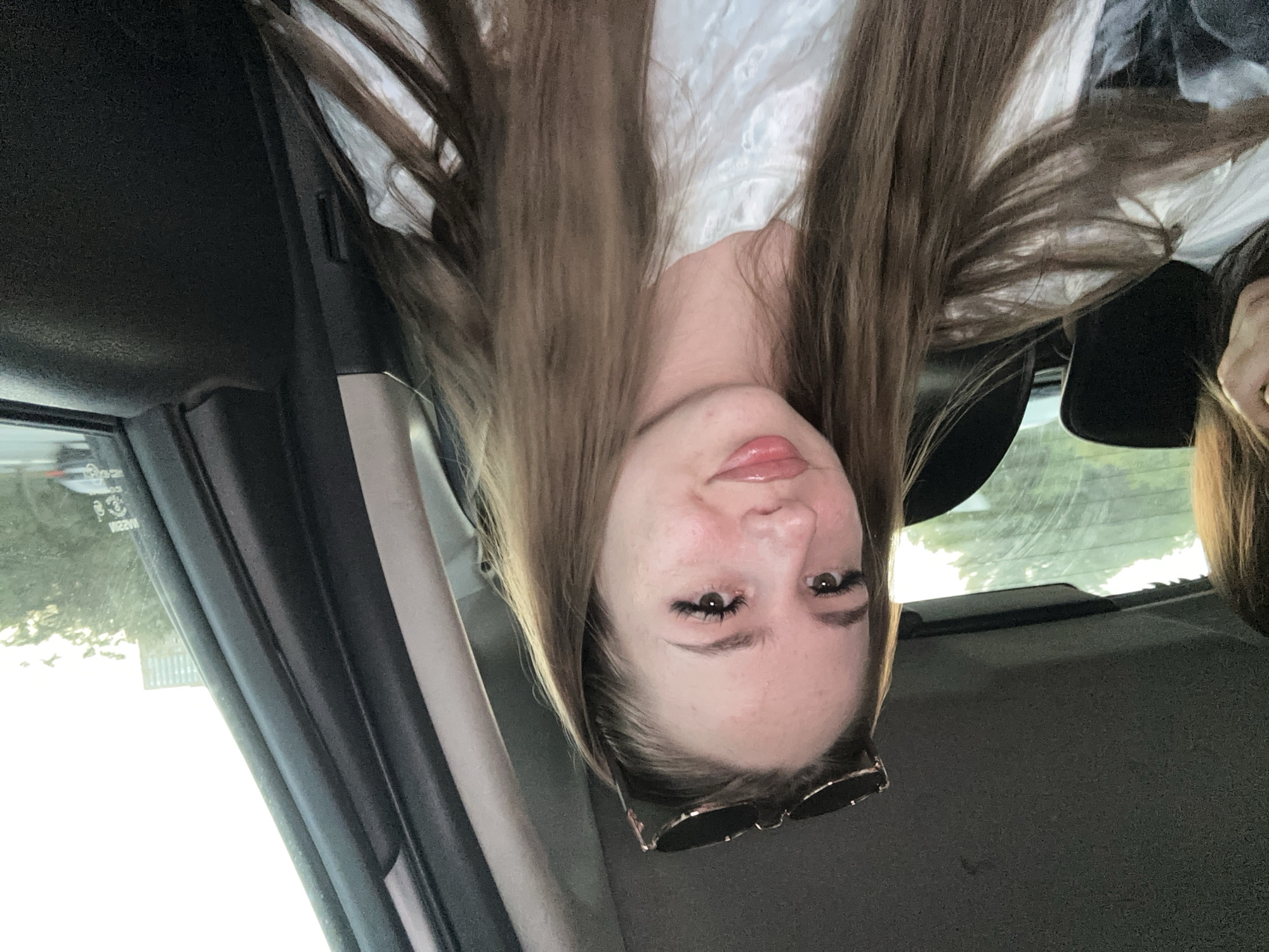 A young woman with long, brown hair and sunglasses on her head is pictured from the chest up, sitting in the backseat of a car. She is wearing a white shirt with a textured pattern. Her eyes are a light brown and she has long eyelashes. Her lips are slightly parted and appear to have a glossy finish. She is looking directly at the camera. The background shows the interior of the car and a blurred view of trees and a building through the window. The scene appears to be in an urban or suburban area.
