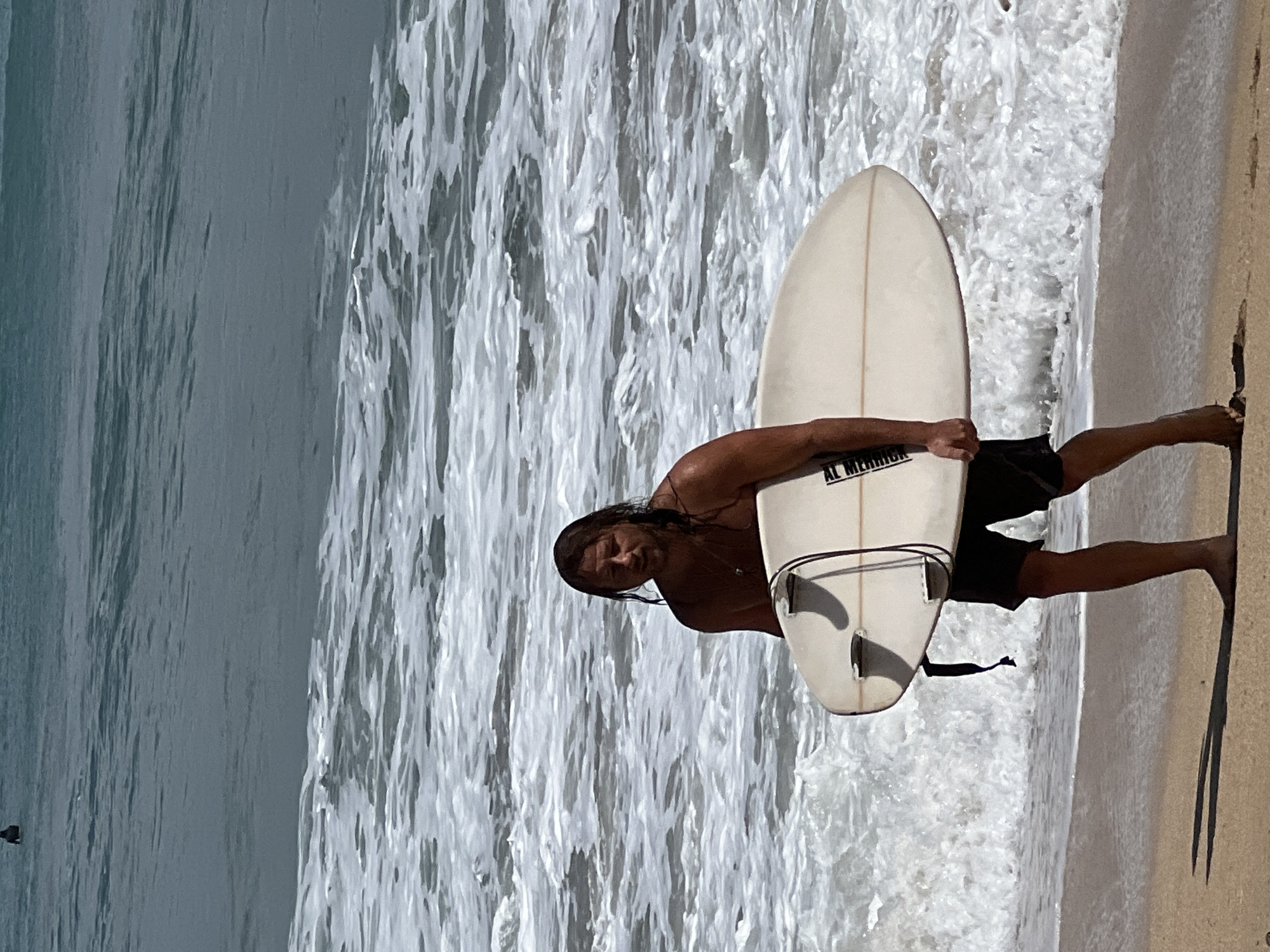 A man with long, wet brown hair is walking on a sandy beach in Kuta, Indonesia, carrying a light-colored surfboard under his right arm. He is wearing black shorts and is shirtless, revealing a tan complexion. The surfboard is oval-shaped with fins. The ocean behind him has moderate waves with white foam crests breaking on the shore. The water is a murky blue-green. The sky is overcast with some patches of sunlight. In the background, another person can be seen in the water. The man is looking towards the camera with a neutral expression. The scene appears to be during the daytime.