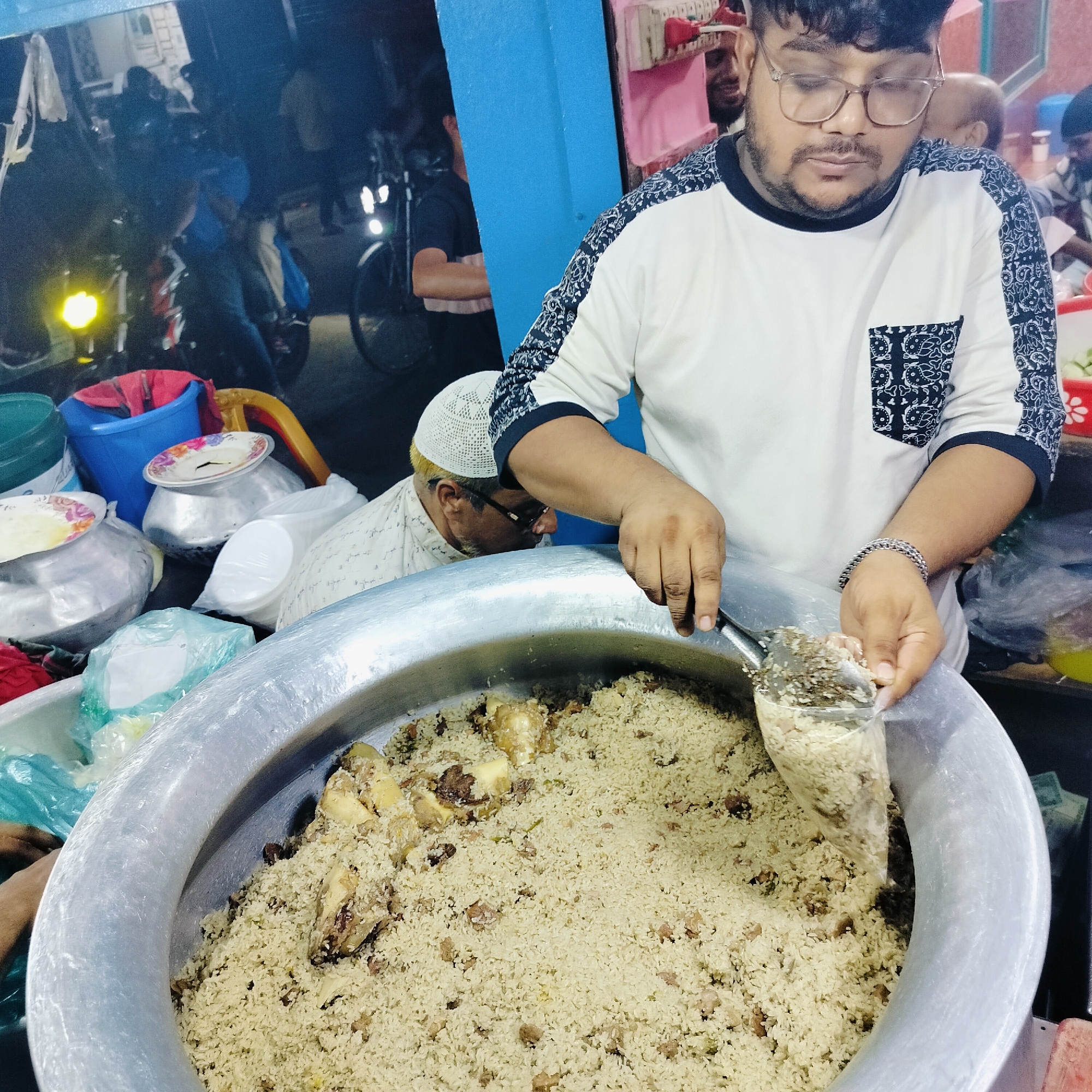 In Dhaka, Bangladesh, a man wearing glasses and a white t-shirt with a black patterned design is serving a large pot of rice and meat with a ladle. He is standing in front of the pot, and another person wearing a white cap and glasses is visible behind him. The scene appears to be an outdoor food stall, with other people and motorcycles in the background. The lighting suggests it is evening or night. The large pot is made of metal and is filled with the food. The man is scooping the food into a clear plastic bag or container.