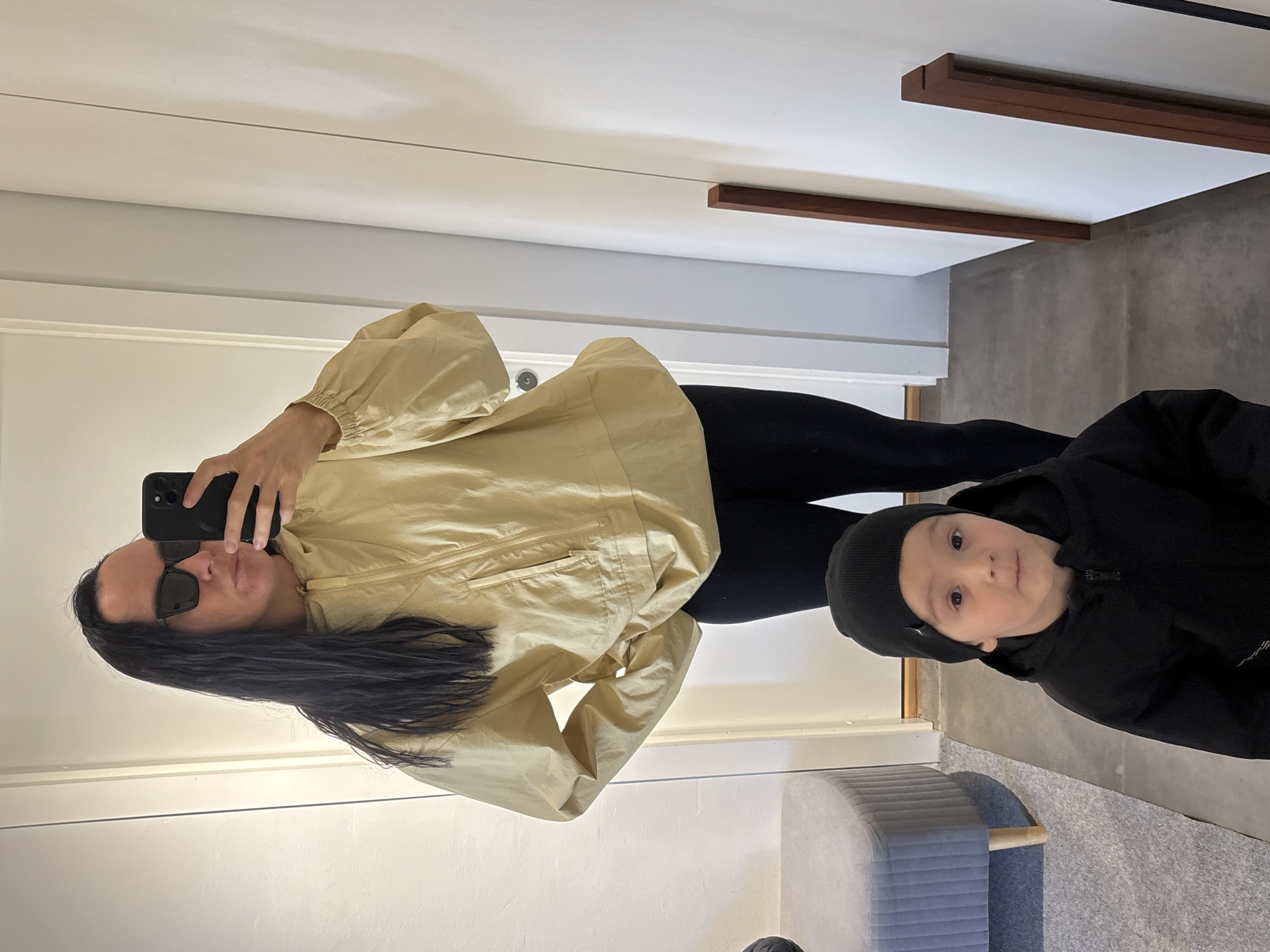 A woman with long black hair and sunglasses is taking a selfie in a mirror. She is wearing a light yellow jacket and black leggings. A young boy in a black beanie and black hooded jacket is standing to her right. The reflection shows part of a room with a gray stool, gray carpet, and white walls. The location is Chaikovsky, Russia.