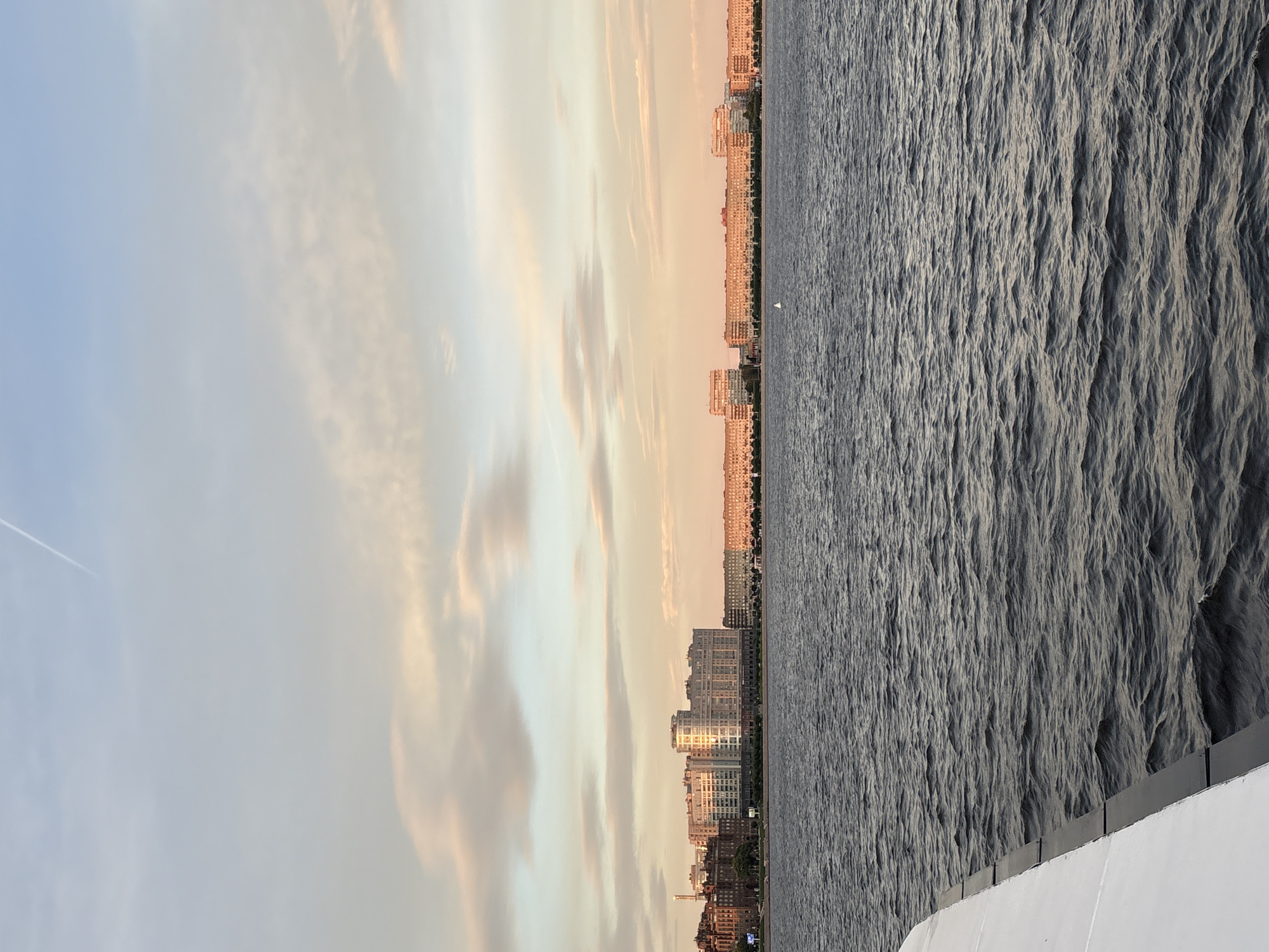 The image shows a wide shot of the Neva River in Saint Petersburg, Russia, at sunset. A portion of a white boat or ferry is visible in the lower left corner. Across the river, a long, low-rise cityscape stretches across the horizon, characterized by apartment buildings with repeating window patterns. The sky above is a gradient of pale blue and soft pink, with scattered wispy clouds and a contrail from an aircraft. The water of the Neva is choppy, with small waves reflecting the warm colors of the sky. The overall impression is of a calm, late evening scene in a major urban setting.