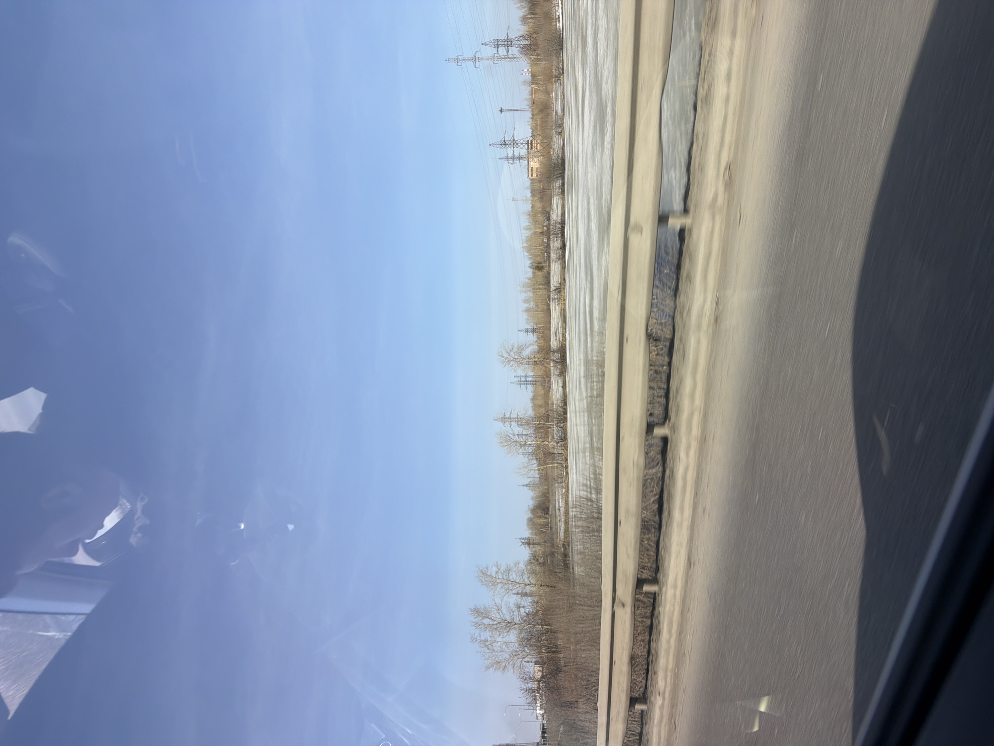 The image depicts a view from a moving vehicle on a highway near Berezniki, Russia. Outside the car window, a roadside barrier runs along the right side of the frame, separating the road from a flat, potentially snowy or icy field with scattered trees and vegetation. In the distance, power lines and industrial structures are visible under a clear, light blue sky. The left side of the image shows the interior of the car, including a partial view of the driver or passenger and part of the car's interior. The road appears to be grey asphalt.