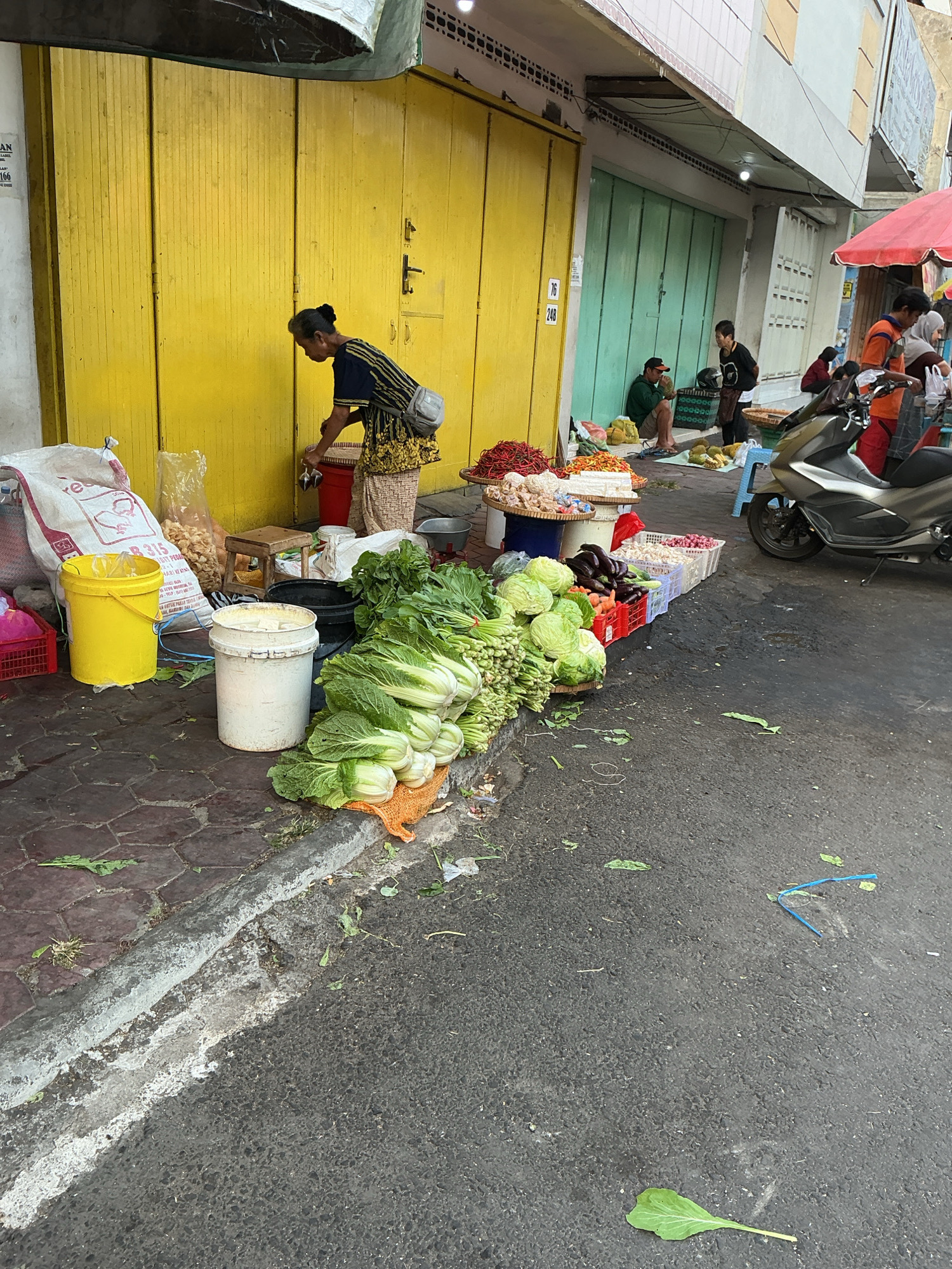 In Magelang, Indonesia, a street scene unfolds with a woman in a patterned skirt and dark shirt standing near a bright yellow storefront. She is tending to a market stall laden with fresh produce, including large heads of green leafy vegetables like napa cabbage and what appear to be bunches of green onions. Beside her are various containers, including a yellow bucket, a white bucket, and baskets. In the background, green doors of shops line the street, and several people are visible, some seated and others standing near a red umbrella and a motorcycle. The ground is paved with cobblestones near the buildings, transitioning to asphalt on the street, with scattered vegetable leaves and debris. The overall atmosphere suggests a bustling local market.