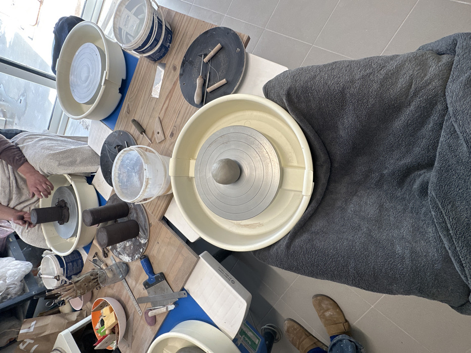 An indoor pottery workshop in Mafra, Portugal, is depicted from a first-person perspective, looking over a dark gray blanket covering the observer's lap. In the immediate foreground, a cream-colored pottery wheel basin holds a metallic wheel head centered with a spherical lump of light gray clay. To the right of this wheel, on a wooden surface, a black circular bat displays three wooden-handled wire cutting tools. The observer's feet, clad in brown boots and blue pants, are visible on a light gray tiled floor to the lower left.
The midground features a wooden tabletop serving as a shared workspace, laden with pottery tools and materials. These include several cylindrical clay forms, some resting on circular bats, and two white plastic buckets, one partially filled. Various hand tools such as a metal scraper with a blue handle, a wooden rib, a metal ruler, carving tools, and sponges are scattered across the surface. A bowl containing colorful sponges and an orange container, along with a container holding numerous modeling tools, are also present.
In the background, a second cream-colored pottery wheel basin is visible on the right. To the upper left, another individual is seated and actively engaged with a third pottery wheel, shaping a cylindrical piece of clay with both hands. This person wears light-colored clothing over a brown long-sleeved shirt. A large window or glass door in the distant background allows natural light into the space, which features a light gray tiled floor throughout. The scene is an active and prepared environment for ceramic production.