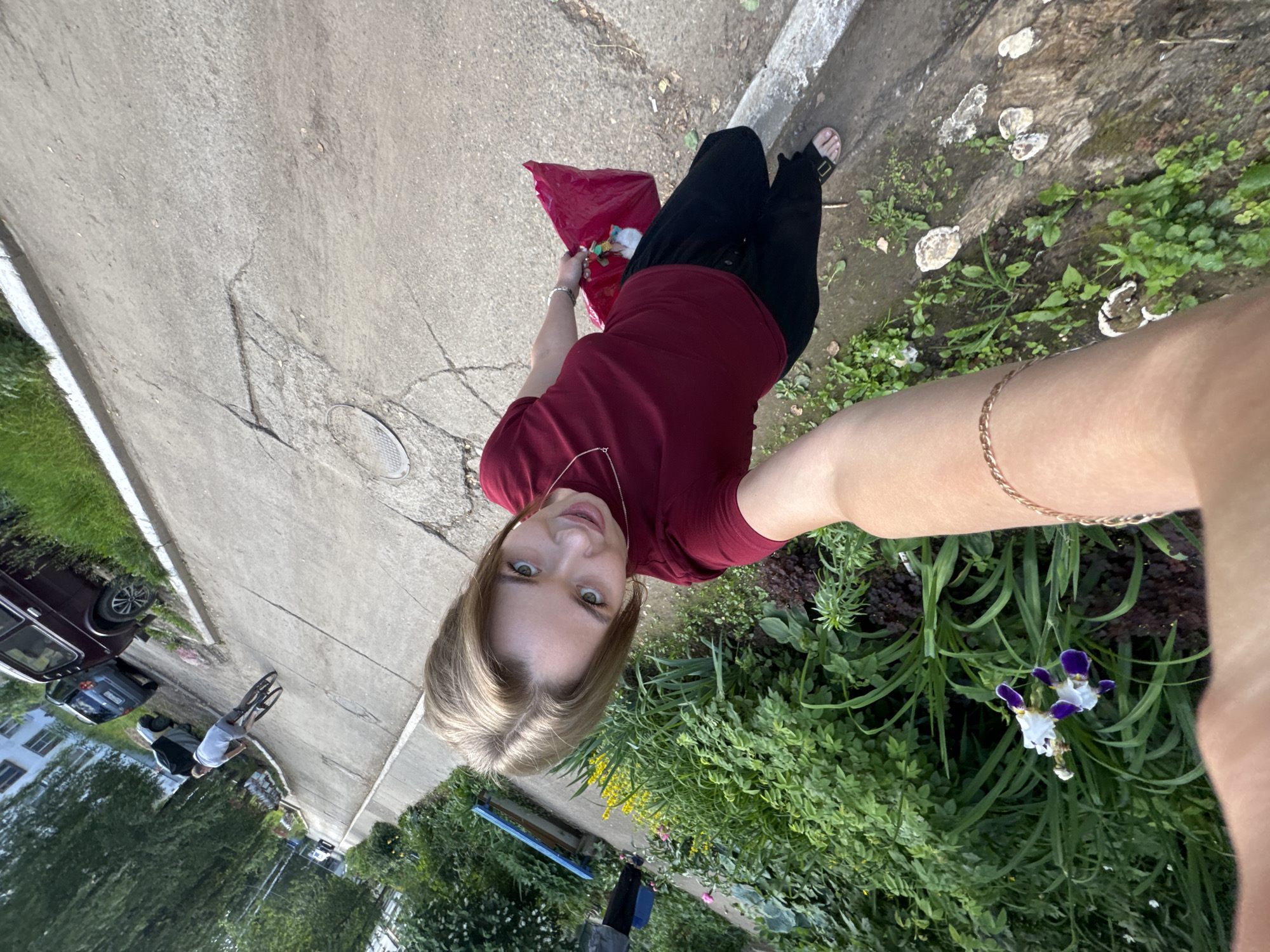 The image is a selfie taken by a young woman with blonde hair and green eyes, wearing a maroon t-shirt and black pants. She is holding a bright pink plastic bag in her right hand. The background shows a paved area, possibly a sidewalk or road, with patches of cracked concrete and grass growing along the edges. Further in the background, there are several parked cars, including a dark-colored Lada. To the left of the woman, there is a flowerbed with green foliage and some purple and white iris flowers. The scene appears to be an outdoor setting in a residential area, likely in the city of Chaikovsky, Russia, as indicated by the presence of Lada cars and the general urban environment. A man on a bicycle is visible in the mid-ground, riding away from the camera. The overall lighting suggests it is daytime.