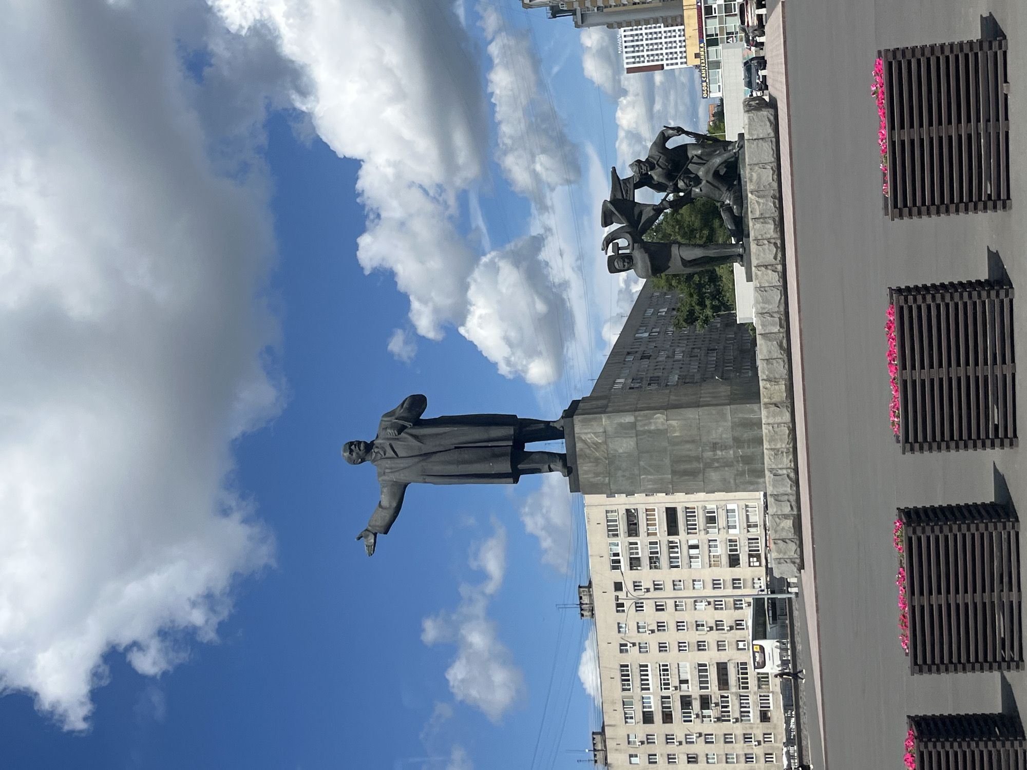An outdoor, ground-level, wide-angle shot captures a monument in a city square on a cloudy day. The monument, centrally located, features a large statue of a man in a coat and hat, with his right arm extended. To the right of the main statue, a smaller group of figures stands, one of whom is raising a flag. The monument is erected on a tall, grey stone pedestal.
Behind the monument, a multi-story, beige apartment building with numerous windows stretches across the background. To the right of this building, a modern, white building with red and grey accents is visible. In the foreground, three dark brown, slatted planters filled with pink flowers are arranged in a line. The sky above is a vibrant blue, dotted with white, fluffy clouds. A few cars are parked on a road in the middle ground, and a person is walking in the distance near the apartment building. The overall scene depicts a public space with a significant historical monument.