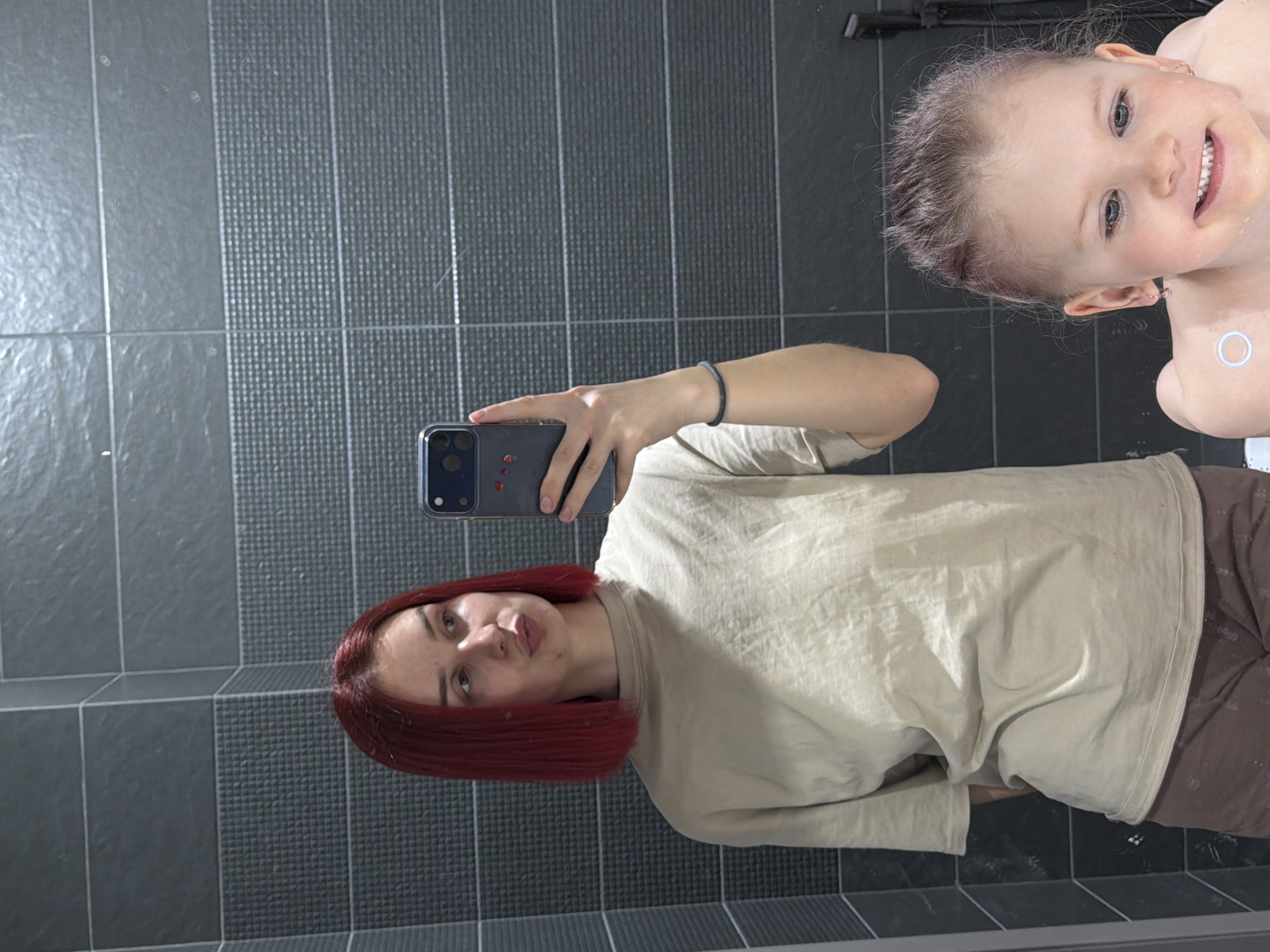A person with red hair is taking a selfie with a child in a bathroom with gray tiled walls. The person is wearing a beige shirt and brown pants. The child is smiling. The location cannot be determined from the image alone.