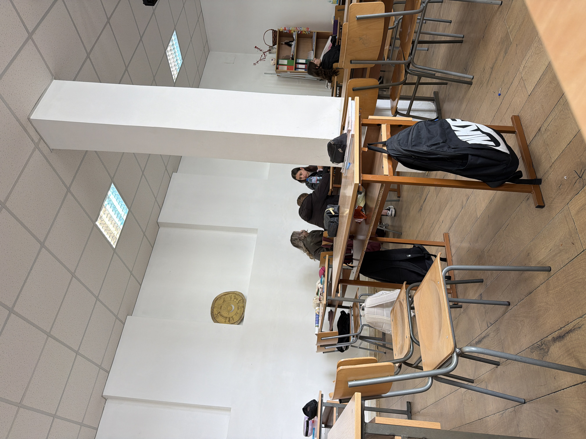 The image shows a classroom in Panagyurishte, Bulgaria. Several rows of desks and chairs are arranged in the room. Students are sitting at the desks, some appearing to be engaged in study or conversation. Backpacks are placed on the floor or hanging on the chairs. A bookshelf is visible at the far end of the room. The walls are white, and a circular decoration is affixed to one of them. The ceiling has a grid pattern with integrated lighting. The floor is made of wooden planks.