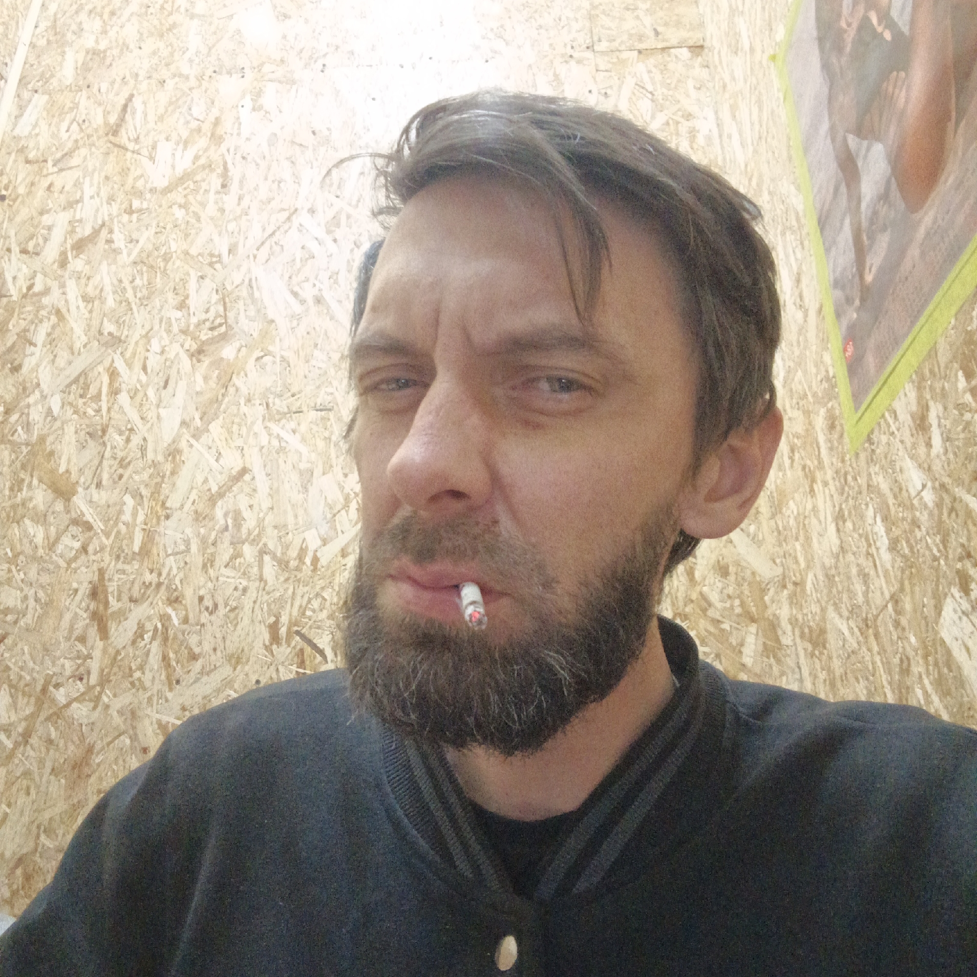 A man with a dark, full beard and brown hair is depicted in a close-up, slightly upward-angled shot. He has a stern and somewhat furrowed expression, with slightly narrowed eyes. A lit cigarette is held between his lips, its glowing tip clearly visible. He is wearing a dark-colored, possibly navy or black, zippered or buttoned jacket or sweatshirt with ribbed details on the collar. The background features walls constructed from Oriented Strand Board (OSB), giving the setting a rustic or industrial feel. To the man's right, a blurry poster or image, framed with green tape, is visible on the OSB wall, showing what appears to be human limbs, specifically legs and feet. The scene takes place in an indoor setting, and the activity shown is the man smoking. The overall impression is candid, taken in Chaikovsky, Russia. No visible text is discernible in the image.