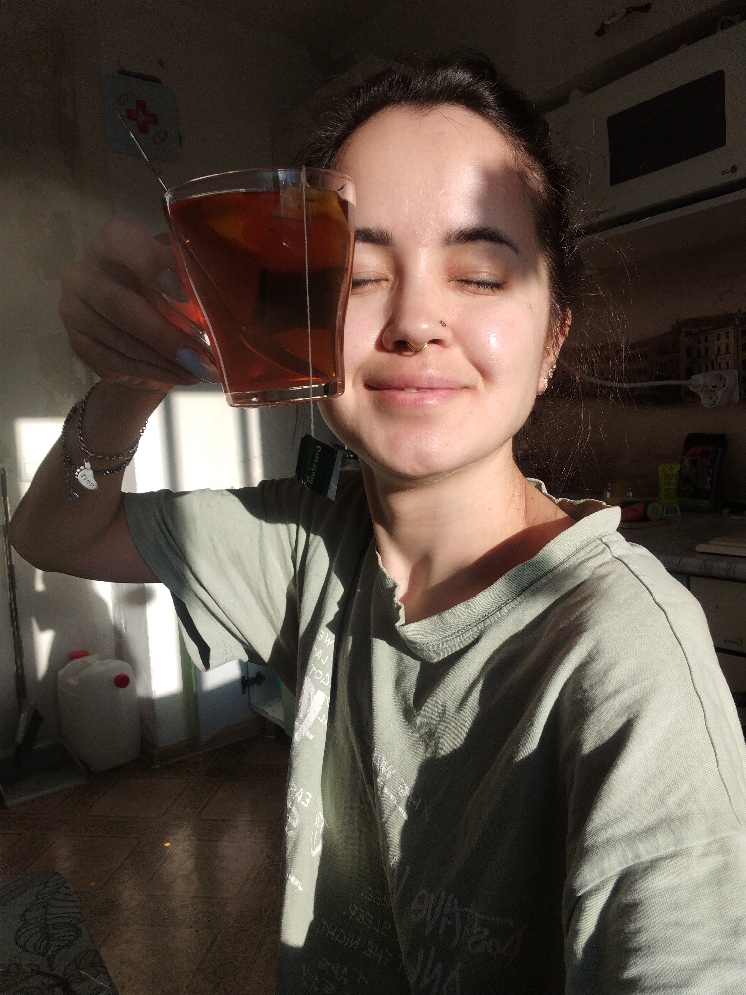 A young woman is depicted indoors, holding a clear glass mug of tea in bright, warm sunlight. She has dark hair, dark eyebrows, and her eyes are closed in a relaxed, smiling expression, conveying contentment. She wears a gold hoop in her septum and a small gold stud on her left nostril. Her light sage green t-shirt has white text printed on it, partially visible and reversed in the shadows, reading phrases like "THE NIGHT", "EVERLASTING", and "BE LOVE". Her left arm is visible, adorned with a silver chain bracelet featuring charms, including a baby footprint charm inscribed with "Virgo". Her fingernails are painted light blue.
The glass mug contains dark amber tea, with a tea bag tag hanging from the side, partially readable as "Greentime". Some fruit slices, possibly lemon or orange, and a spoon are visible in the tea.
The setting appears to be a domestic interior, likely a kitchen or a utility area, illuminated by strong natural light streaming from an unseen window. This light casts sharp shadows across the scene, indicating either early morning or late afternoon. In the background, a white LG microwave oven is mounted above a counter or shelf. Below, a white plastic canister with red caps, a dustpan and broom, and a power strip are visible. The floor has light-colored patterned tiles or linoleum. The overall ambiance is peaceful and warm, highlighted by the golden light.