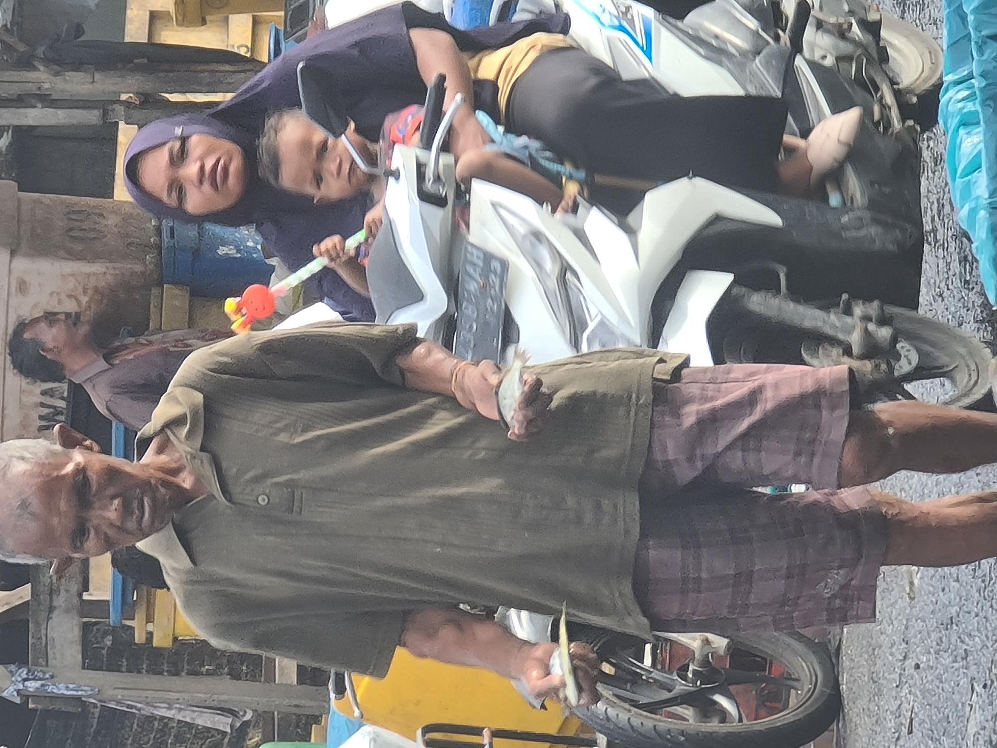An outdoor scene in Lhokseumawe, Indonesia, shows three people in the foreground with a motorcycle. In the left foreground, an elderly man stands wearing a dark olive green short-sleeved collared shirt and plaid shorts. He is holding two small fish, one in each hand. To his right, a woman wearing a dark purple hijab and dark clothing is seated on a white motorcycle. She is holding a young child, who is facing the camera and holding a colorful toy. The child is wearing a brightly colored shirt. The motorcycle is white with blue accents and has a license plate. In the background, a man wearing a light brown collared shirt is partially visible. The environment appears to be a marketplace or street, with yellow bins and structures in the background. The ground is paved with gravel.
