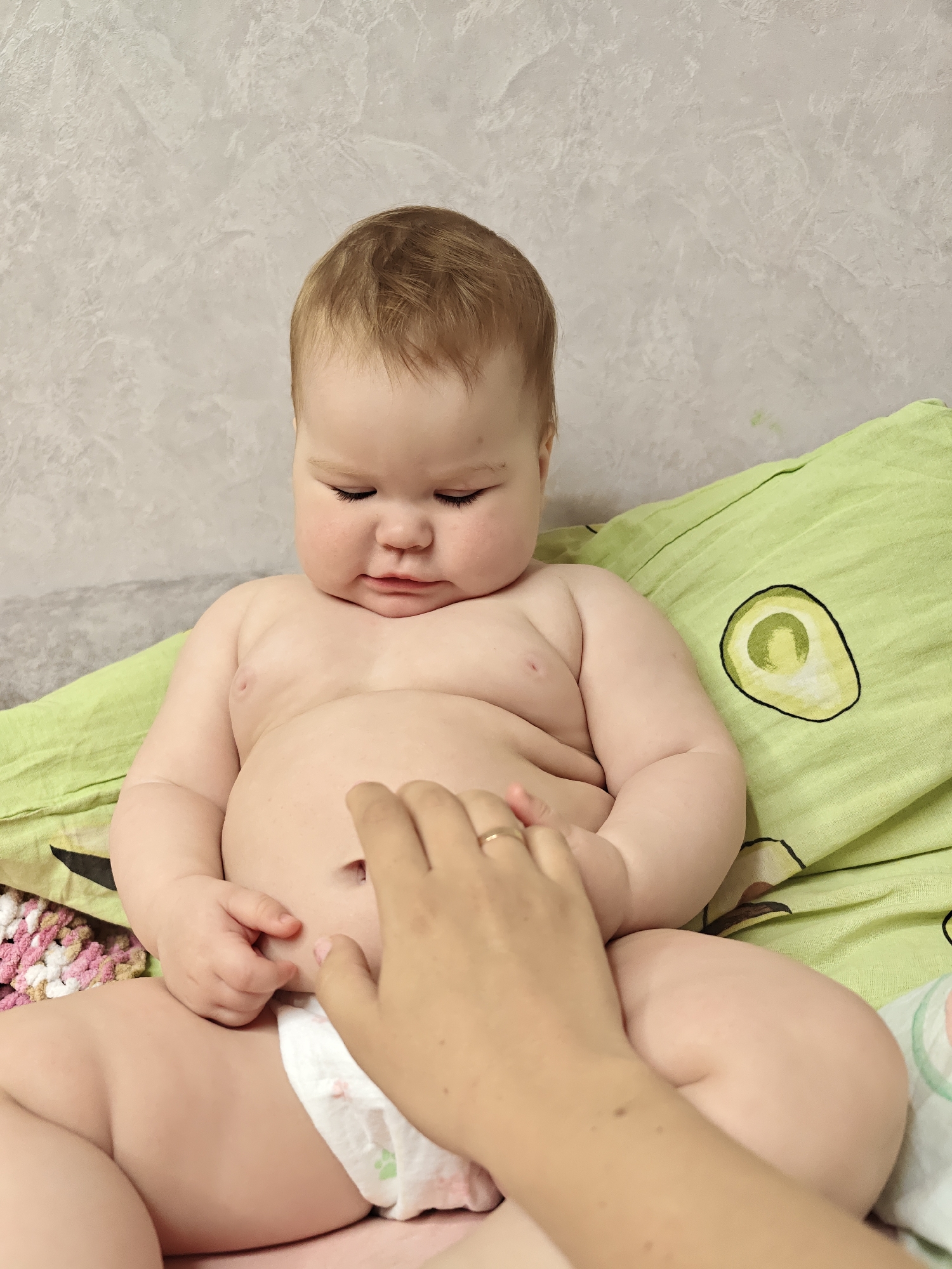 A baby sits on a bed, wearing a diaper. A hand, likely belonging to an adult, touches the baby's belly. The bed has a green avocado-patterned pillow and a crocheted blanket. The background is a light-colored wall. The image was taken in Chaikovsky, Russia.