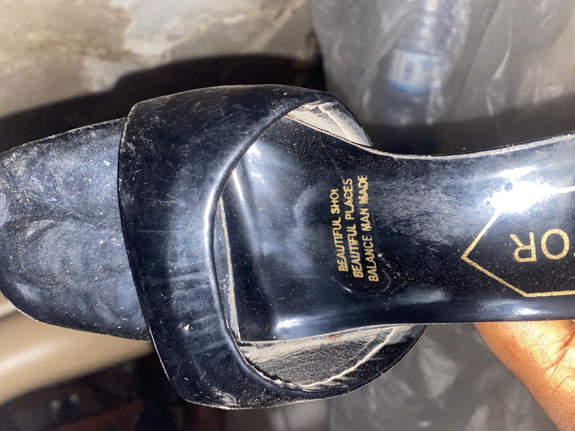 The image shows a close-up of a black sandal with gold lettering on the sole. The lettering reads "BEAUTIFUL SHOE", "BEAUTIFUL PLACES", and "BALANCE MAN MADE". The sandal appears to be dusty. A hand is visible in the lower left corner, holding the sandal. The background is blurred and appears to be an indoor setting with some plastic-wrapped items. The location in the city of Jauro Yinu, Nigeria is not discernible from the image.