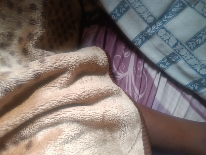 The scene is located in Oye, Nigeria. The image captures a close-up view of an indoor setting, likely a bed. A dark-skinned human forearm is visible on the right side, extending from beneath a light brown, plush blanket with darker brown spots, resembling a leopard print pattern. The blanket appears rumpled. Behind the arm and blanket, two distinct fabric patterns are present: an upper right section displays a light blue fabric with a white floral and grid motif, while a lower background section features a purple fabric with prominent white swirling patterns. The overall impression is of a person resting under bedding. No faces or specific actions beyond resting are discernible.